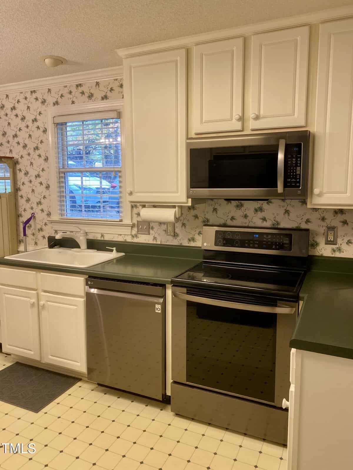 A kitchen with white cabinets, green countertops, stainless steel appliances including a stove, microwave, and dishwasher, a sink under a window, and wallpaper with a floral pattern. The floor is light-colored with a square tile pattern.