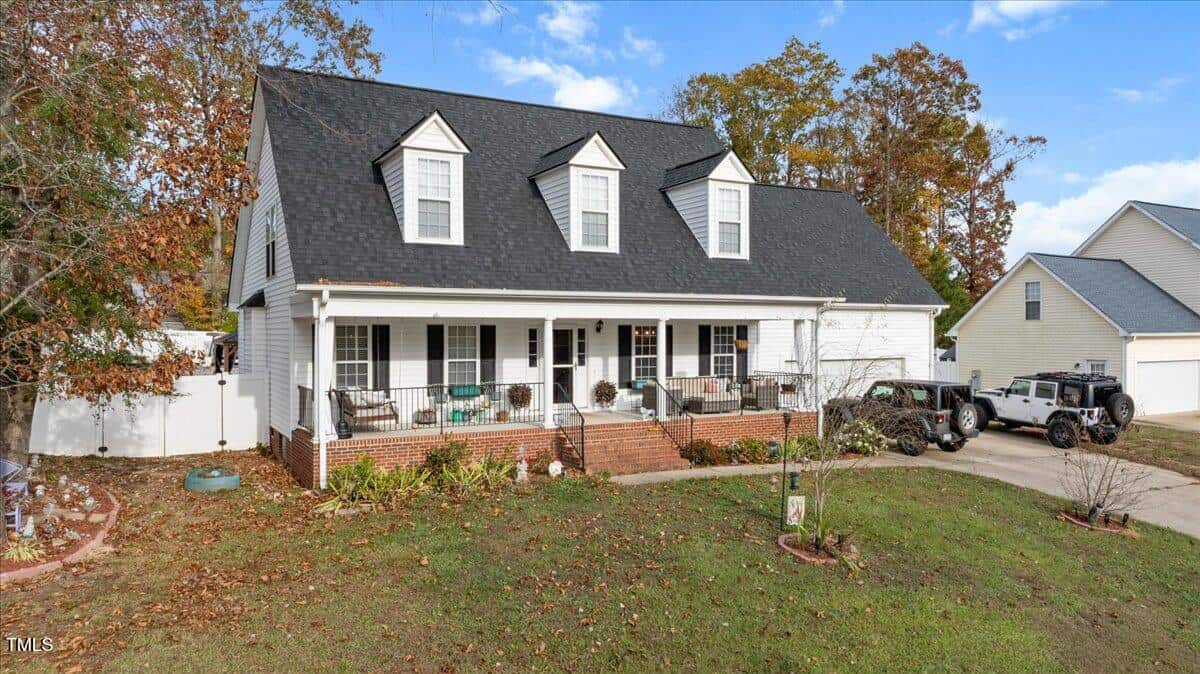 A two-story house with black roof shingles, white siding, and a covered front porch with seating. The front yard has light landscaping, driveway, and two parked vehicles. Trees with autumn leaves are in the background.
