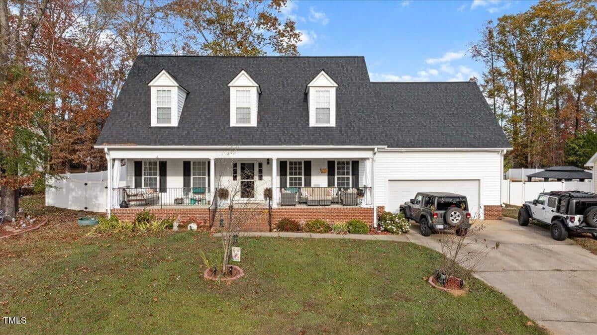 A two-story white house with three dormer windows, a covered front porch, and a black roof. Two vehicles are parked in the driveway, and there is a lawn with some scattered leaves in front. Trees border the property.