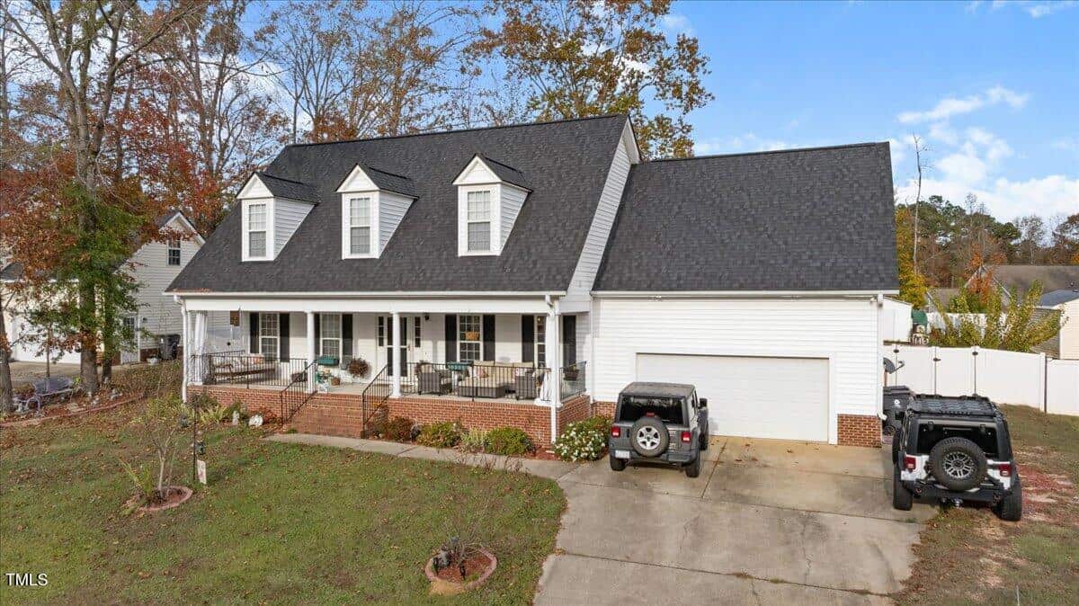 A white two-story house with three dormer windows, a front porch, and a two-car garage. Two vehicles are parked in the driveway, surrounded by grass and trees. The sky is blue with some clouds.