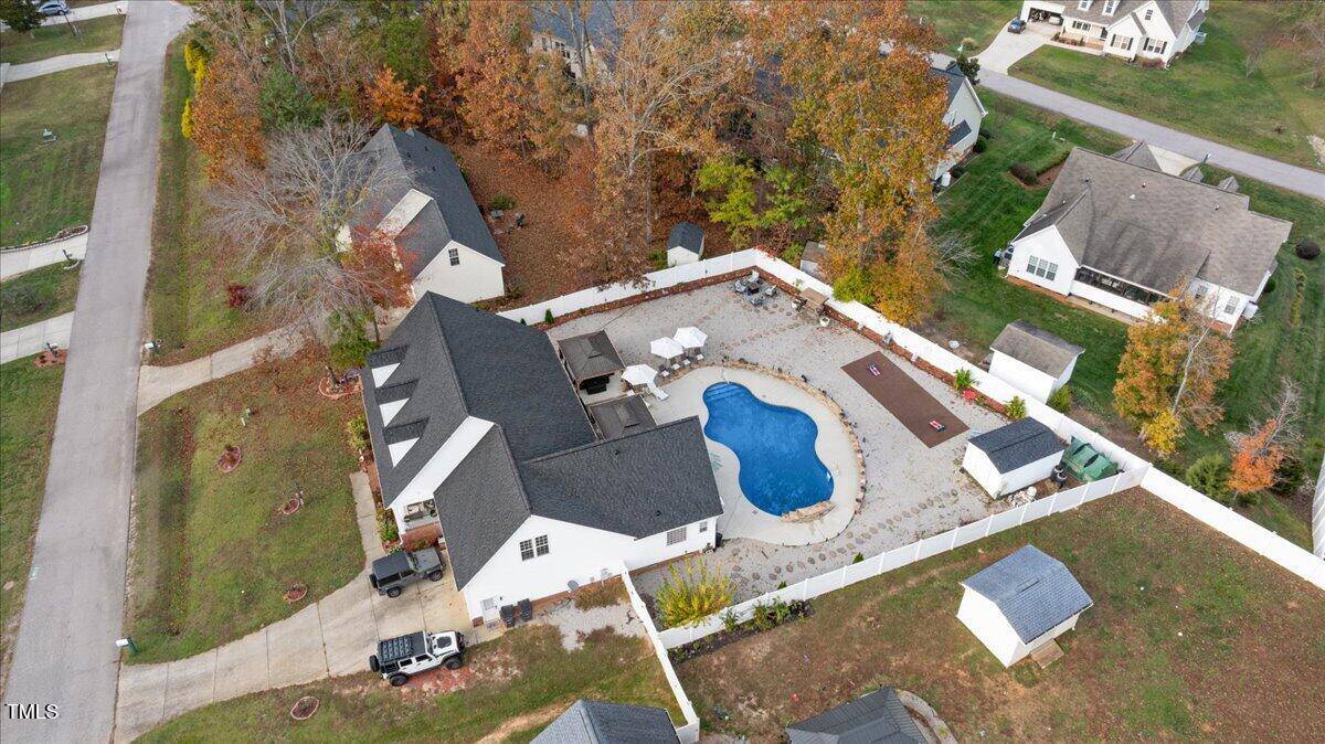Aerial view of a suburban house with a fenced backyard featuring an irregularly-shaped swimming pool, patio furniture, and surrounding landscaping. Nearby houses and trees are also visible.