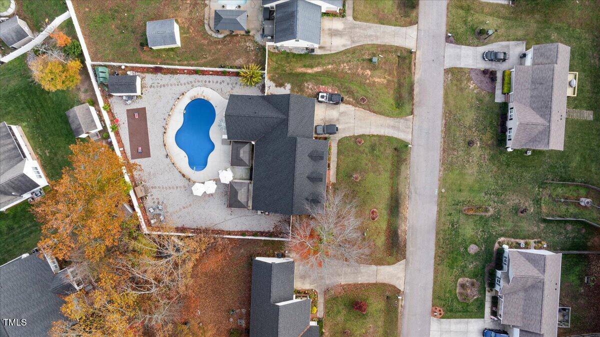 Aerial view of a suburban house with a fenced backyard, patio, and a kidney-shaped swimming pool. Surrounding homes, lawns, driveways, and a street are also visible.