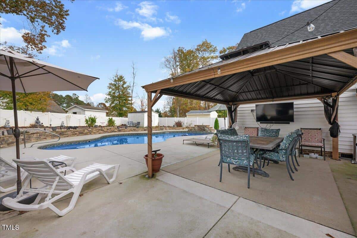 Covered patio with dining table and chairs next to a pool. Lounge chairs, umbrella, potted plants, and a TV visible. Trees and fence in background under a partly cloudy sky.