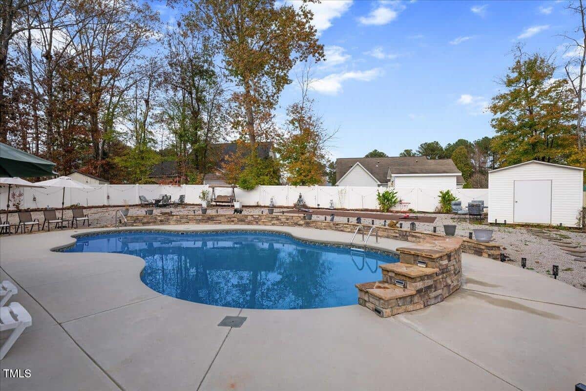 A backyard with an in-ground, kidney-shaped swimming pool surrounded by a concrete deck, stone steps, white fence, and trees. There are garages and a gravel area in the background.