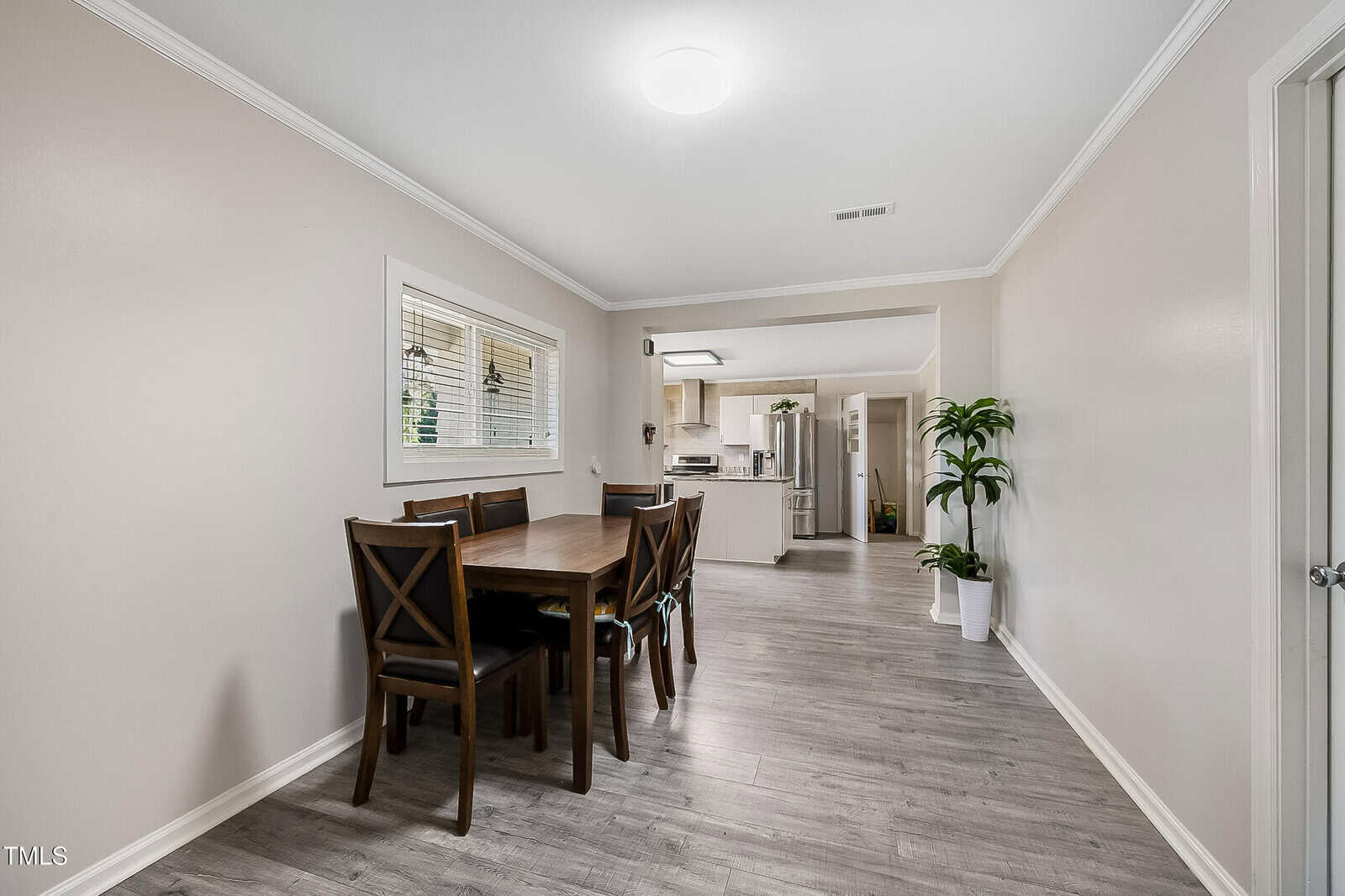 A dining room with a wooden table and six matching chairs on a light gray wood floor. A window with blinds is on the left wall. Two potted plants are in the background near the entrance to the kitchen, which has white cabinets and appliances.