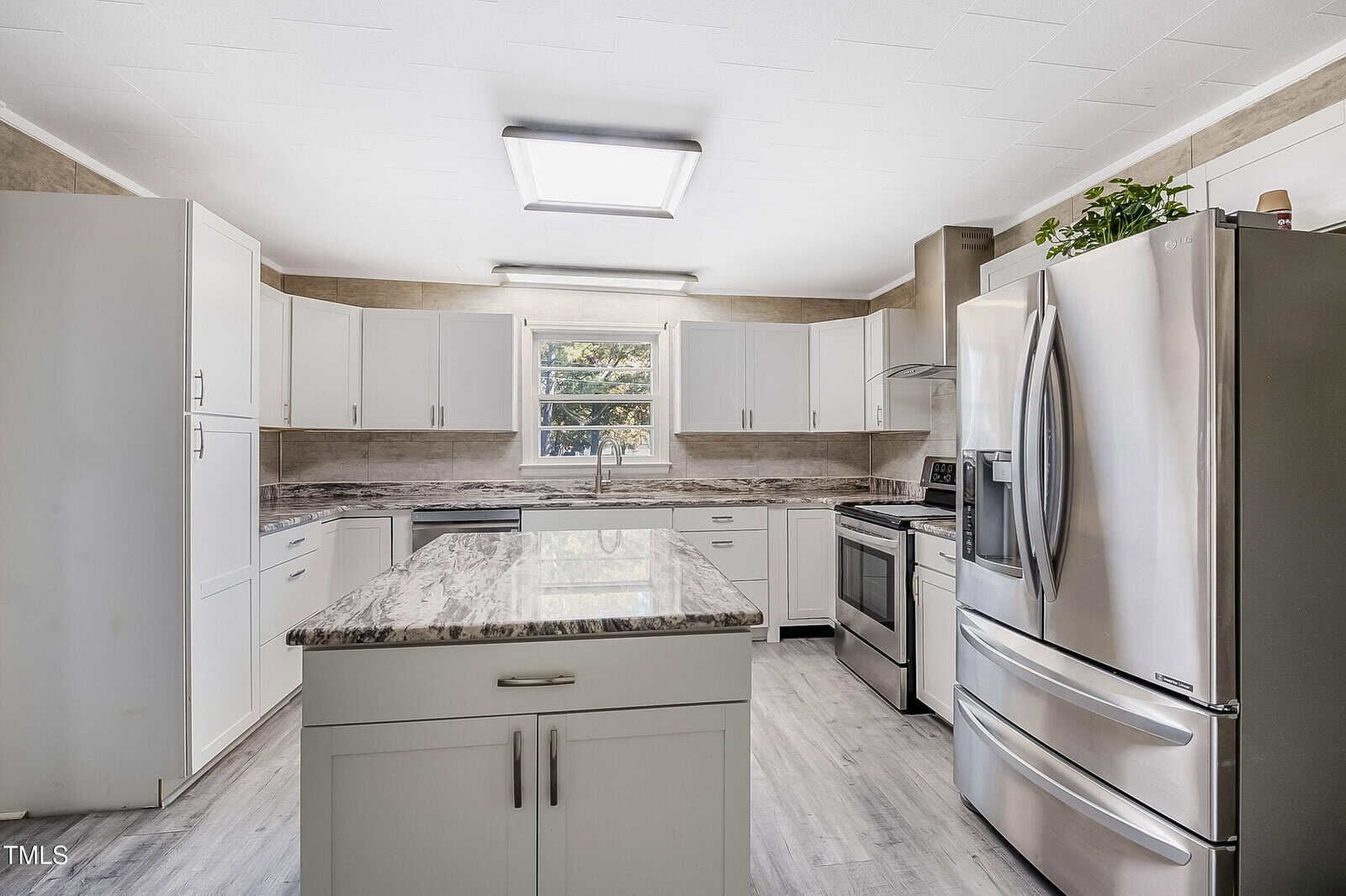 A modern kitchen with white cabinets, stainless steel appliances, and a central island with a marble countertop. Natural light enters through a window above the sink. The floor is light-colored hardwood.