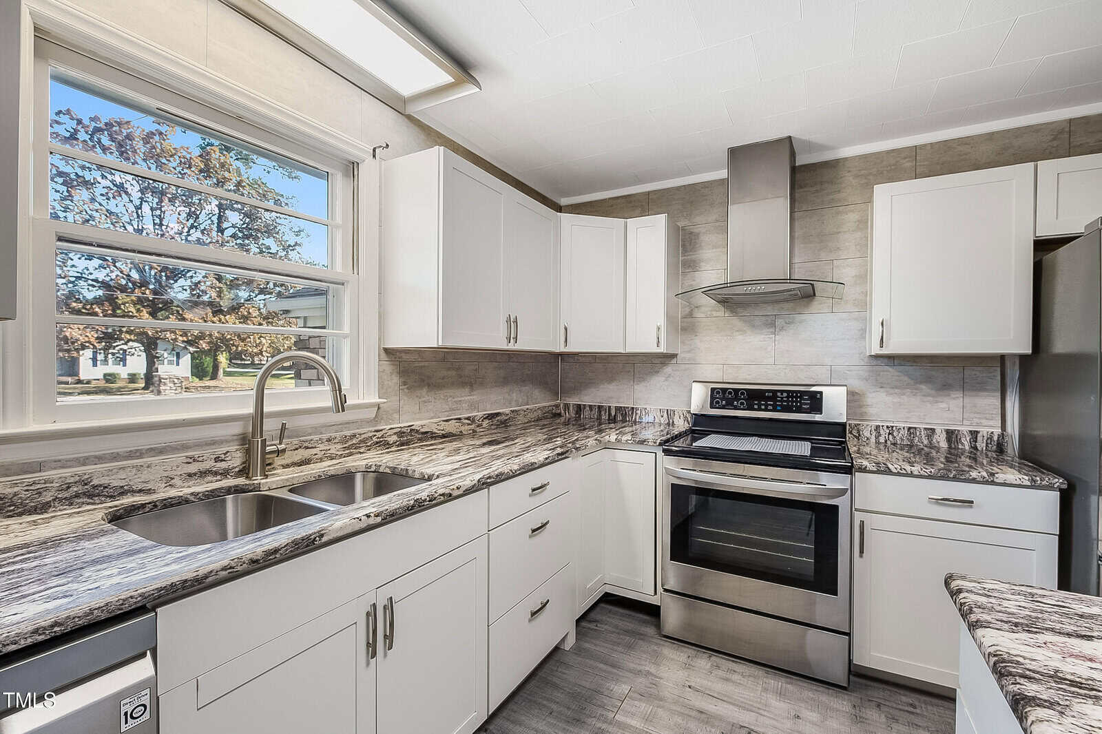 Modern kitchen with white cabinets, stainless steel appliances, and a double sink under a large window. The countertops are dark with a marbled pattern. A range hood is above the stove, and gray tile flooring complements the neutral color scheme.