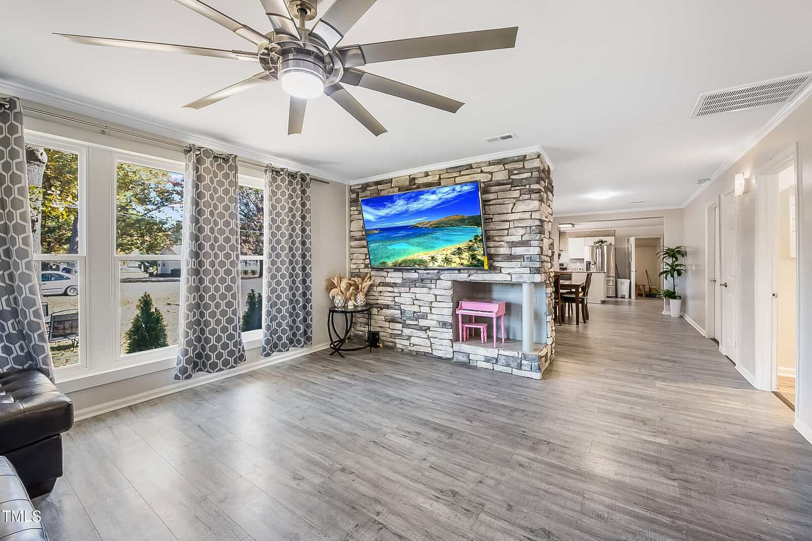 A living room with light wood flooring and a stone fireplace featuring a flat-screen TV displaying a beach scene. Large windows with patterned curtains allow natural light. A ceiling fan is in view, with a corridor leading to other rooms.