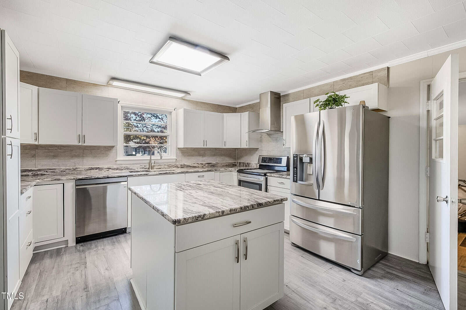 A modern kitchen with white cabinets and stainless steel appliances, including a refrigerator and dishwasher. The center features a large island with a granite countertop. A plant sits atop the cabinets, and a window provides natural light.