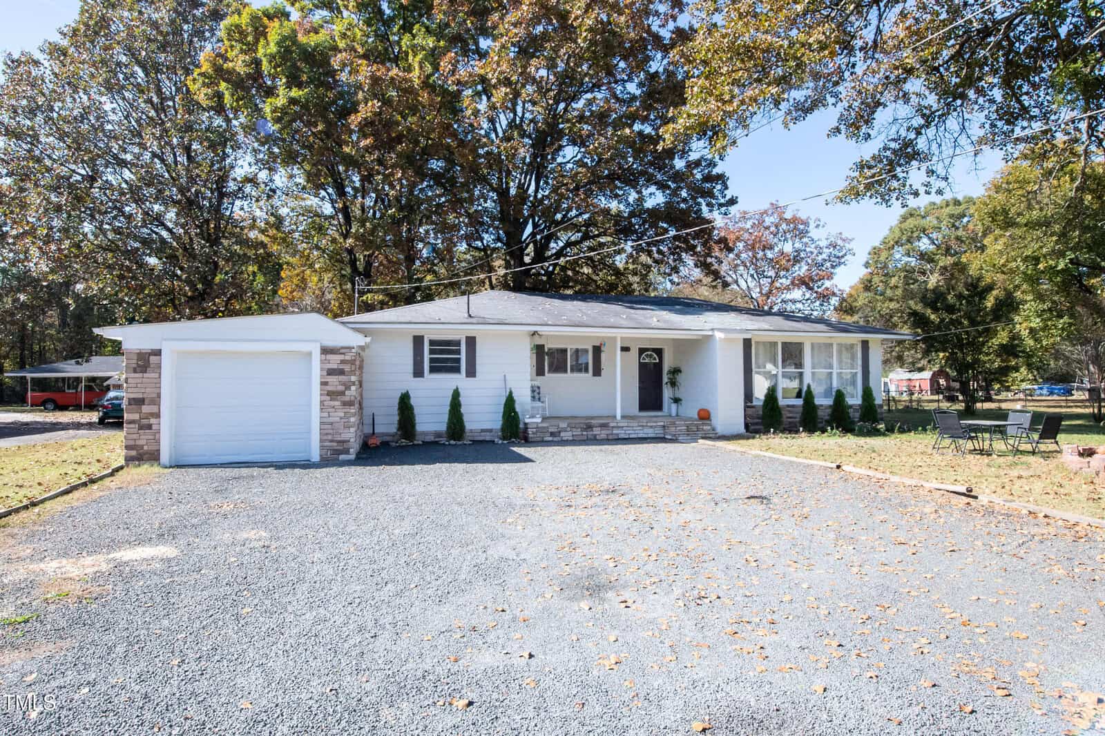 A single-story white house with a garage and a gravel driveway. The yard has scattered autumn leaves, small shrubs along the front, and a small patio area with outdoor furniture. Trees with some remaining leaves border the property.