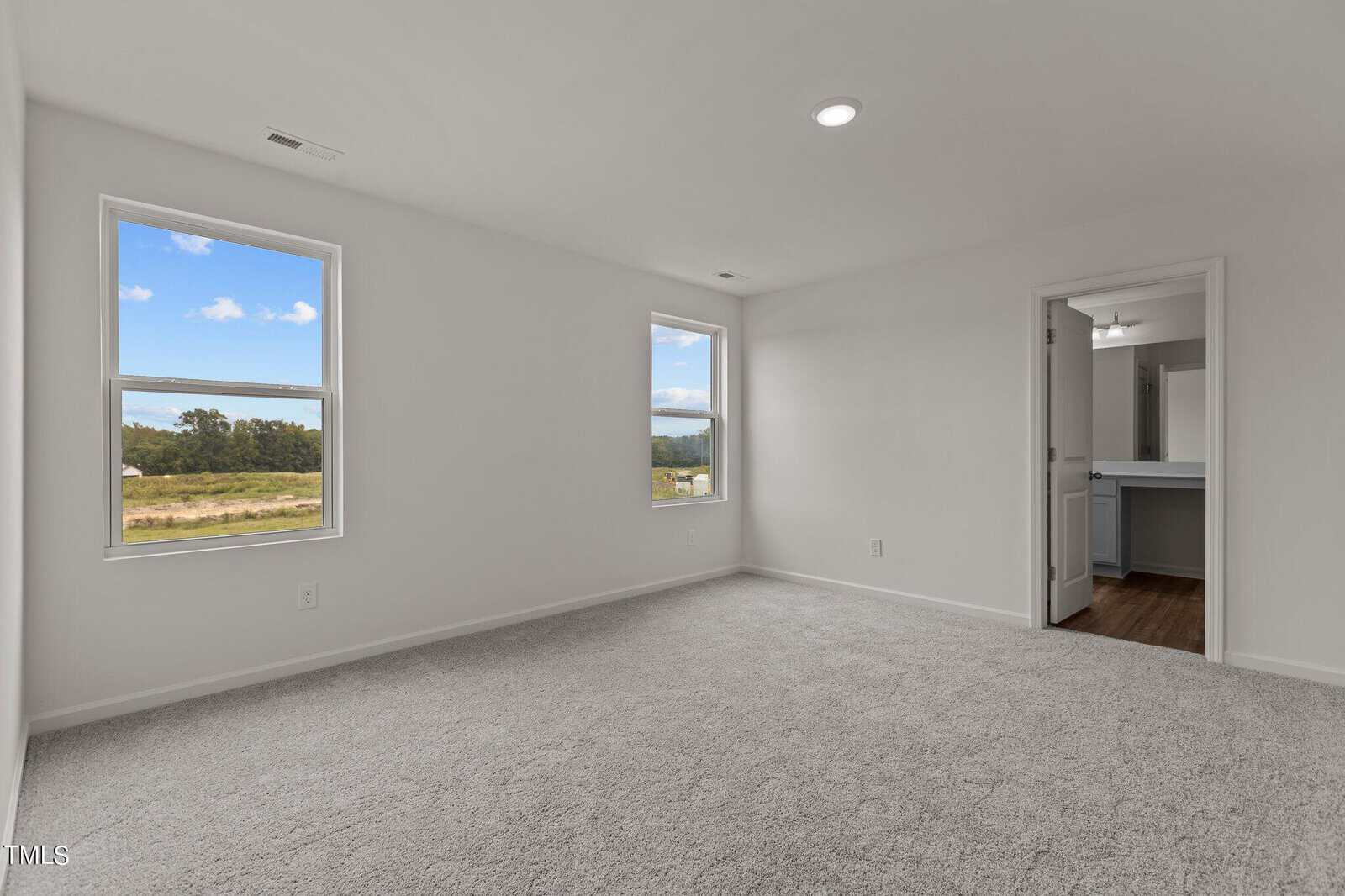 Bright, empty bedroom with light gray carpet, two windows with outdoor views, white walls, and an open doorway leading to a bathroom with a vanity and mirror.