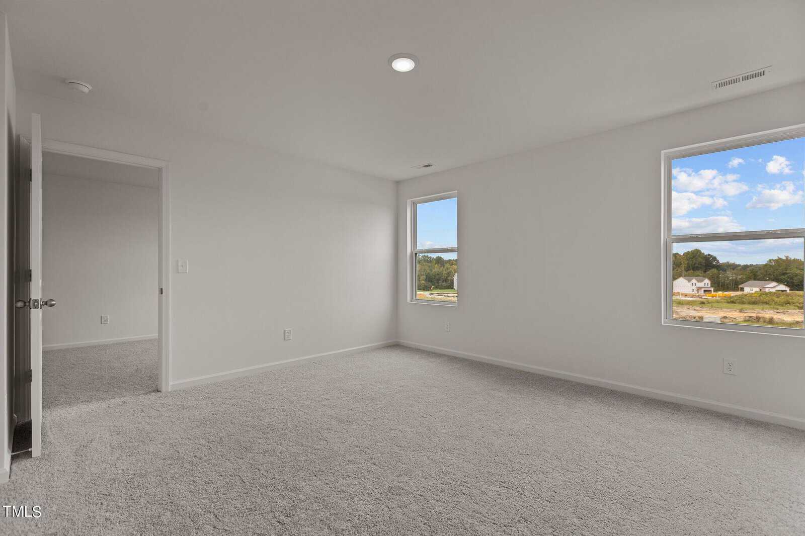 Empty bedroom with light gray carpet, white walls, and two large windows showing a clear sky and outdoor scenery. An open door on the left leads to another room. Ceiling light is turned on.