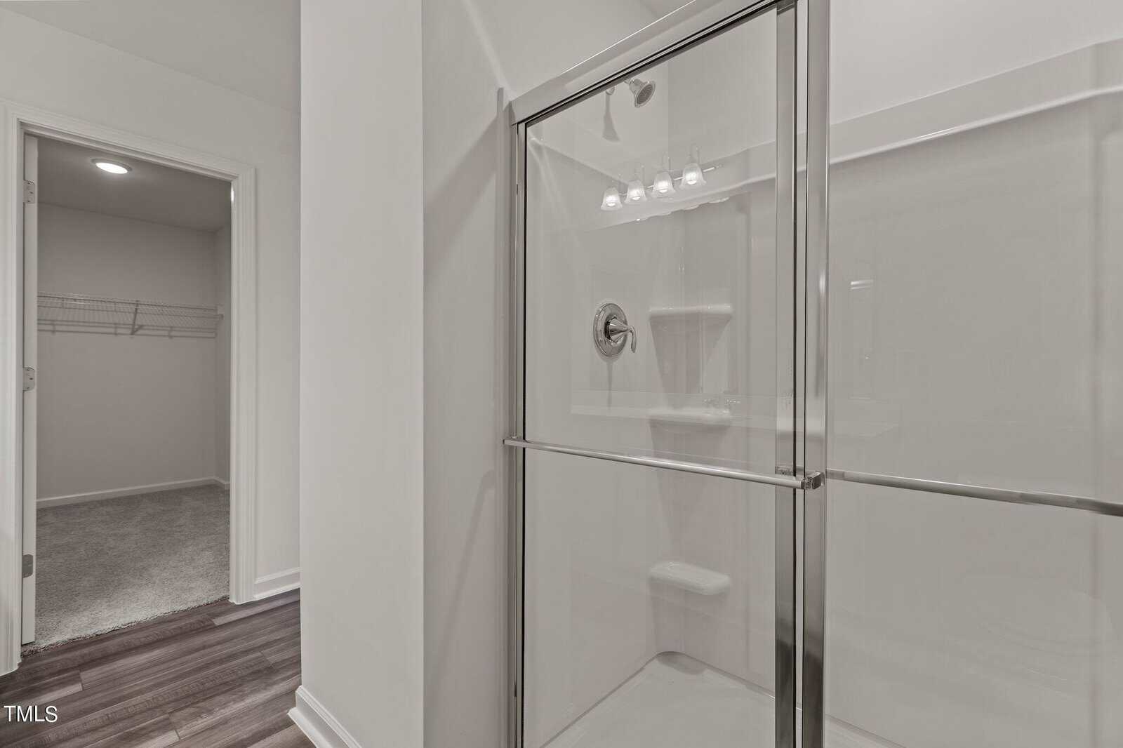 A glass shower with sliding doors is next to a doorway leading to a carpeted walk-in closet with wire shelving. The bathroom has white walls and dark wood flooring.