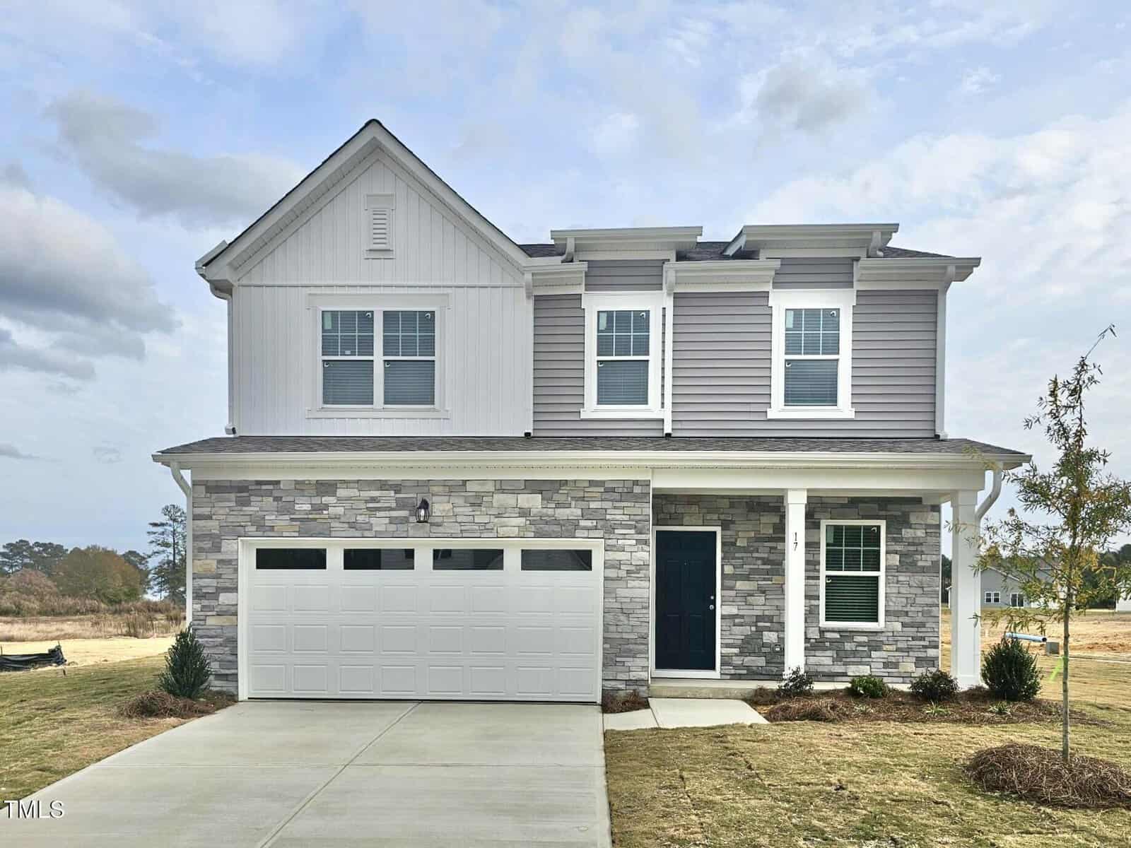 Two-story suburban house with a two-car garage, light gray stone and siding exterior, covered front porch, and a small front yard with new landscaping under a partly cloudy sky.