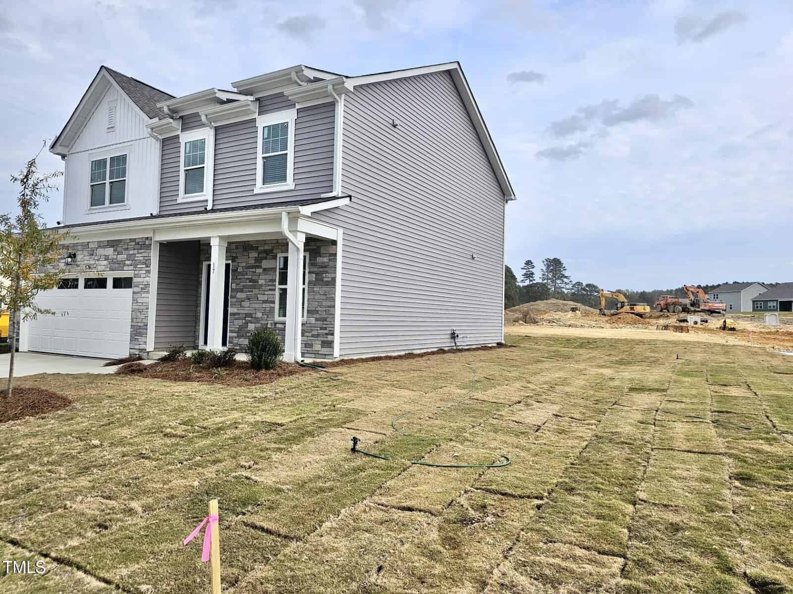 A newly constructed two-story house with gray siding and stone accents stands next to a yard with freshly laid sod. Construction equipment and unfinished lots are visible in the background.
