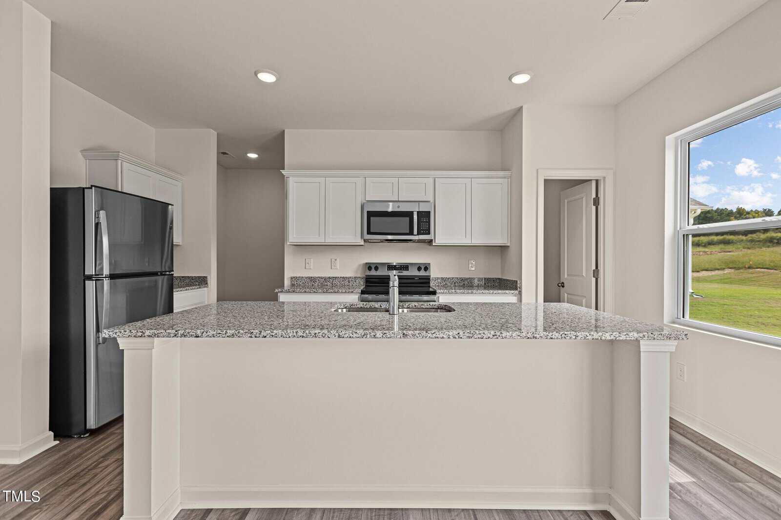 Modern kitchen with white cabinets, granite countertops, stainless steel appliances, and a large center island. Sunlight streams through a window showing a green lawn and blue sky outside.