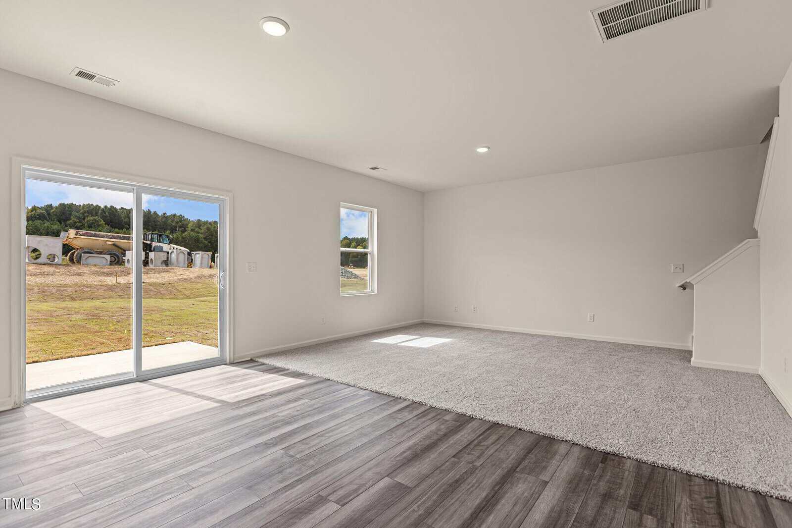 Spacious, empty living room with light gray carpet and wood flooring, white walls, two windows, sliding glass door leading outside, and a staircase on the right. Construction equipment visible through the door.
