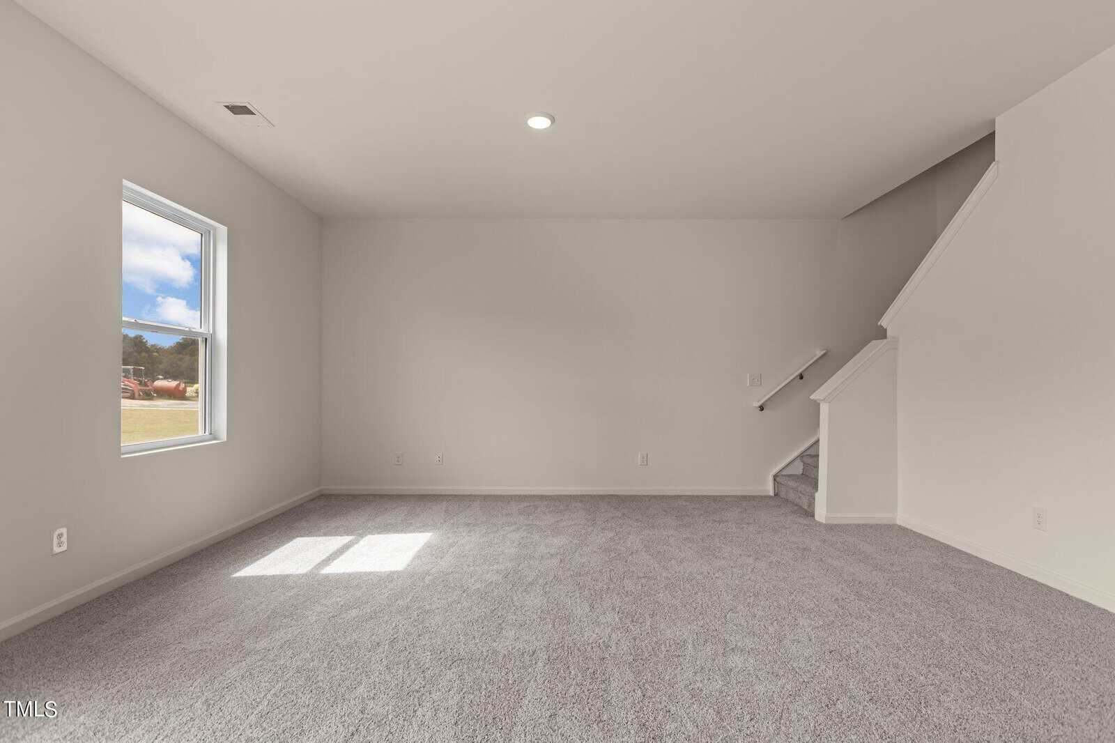 A spacious, empty room with light gray carpet, white walls, a single window letting in daylight, and a staircase in the corner leading upstairs.