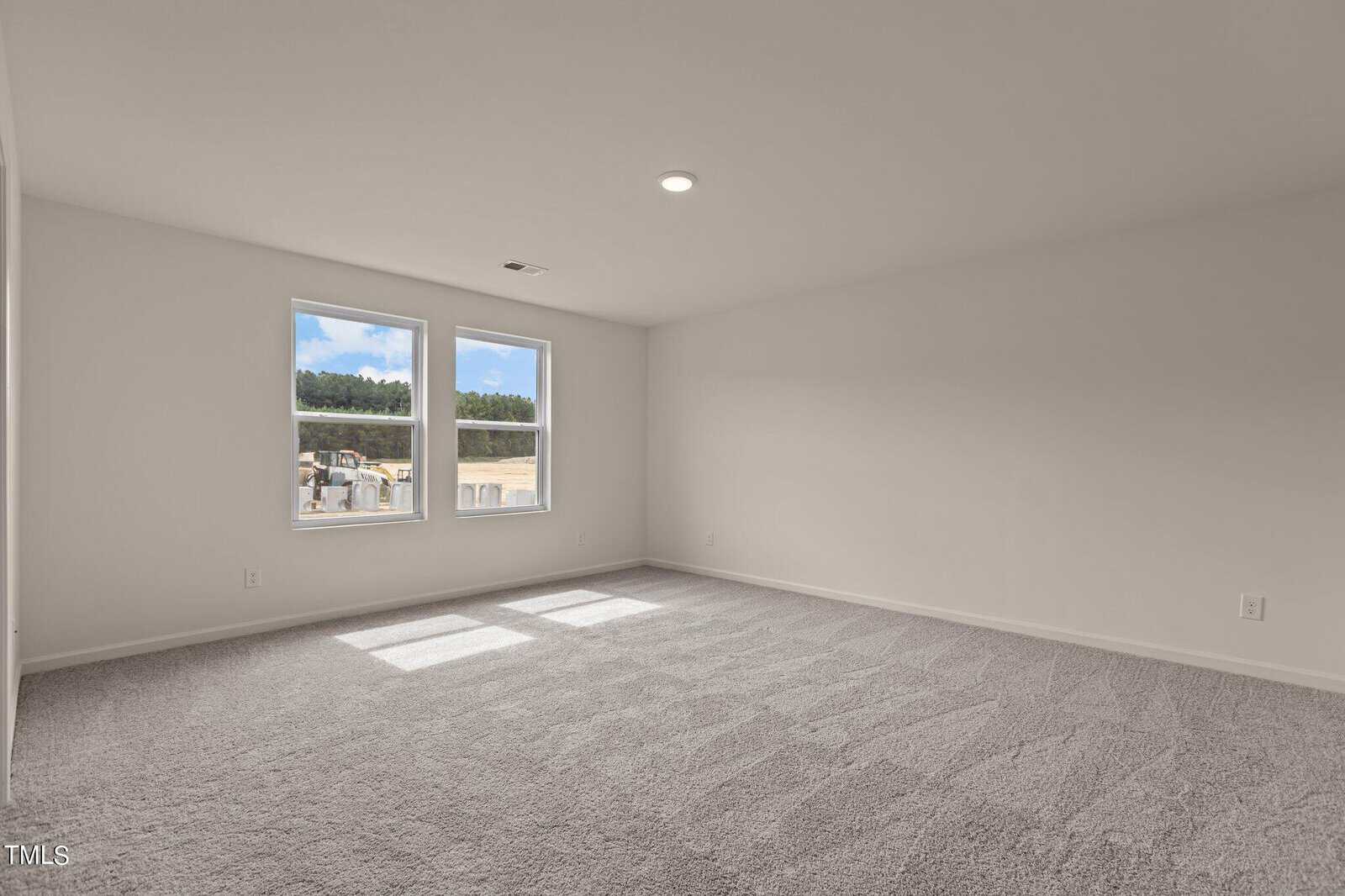Spacious, empty room with light gray carpet, white walls, and two large windows letting in natural light. View outside shows construction and greenery under a blue sky. Ceiling light and air vent visible.