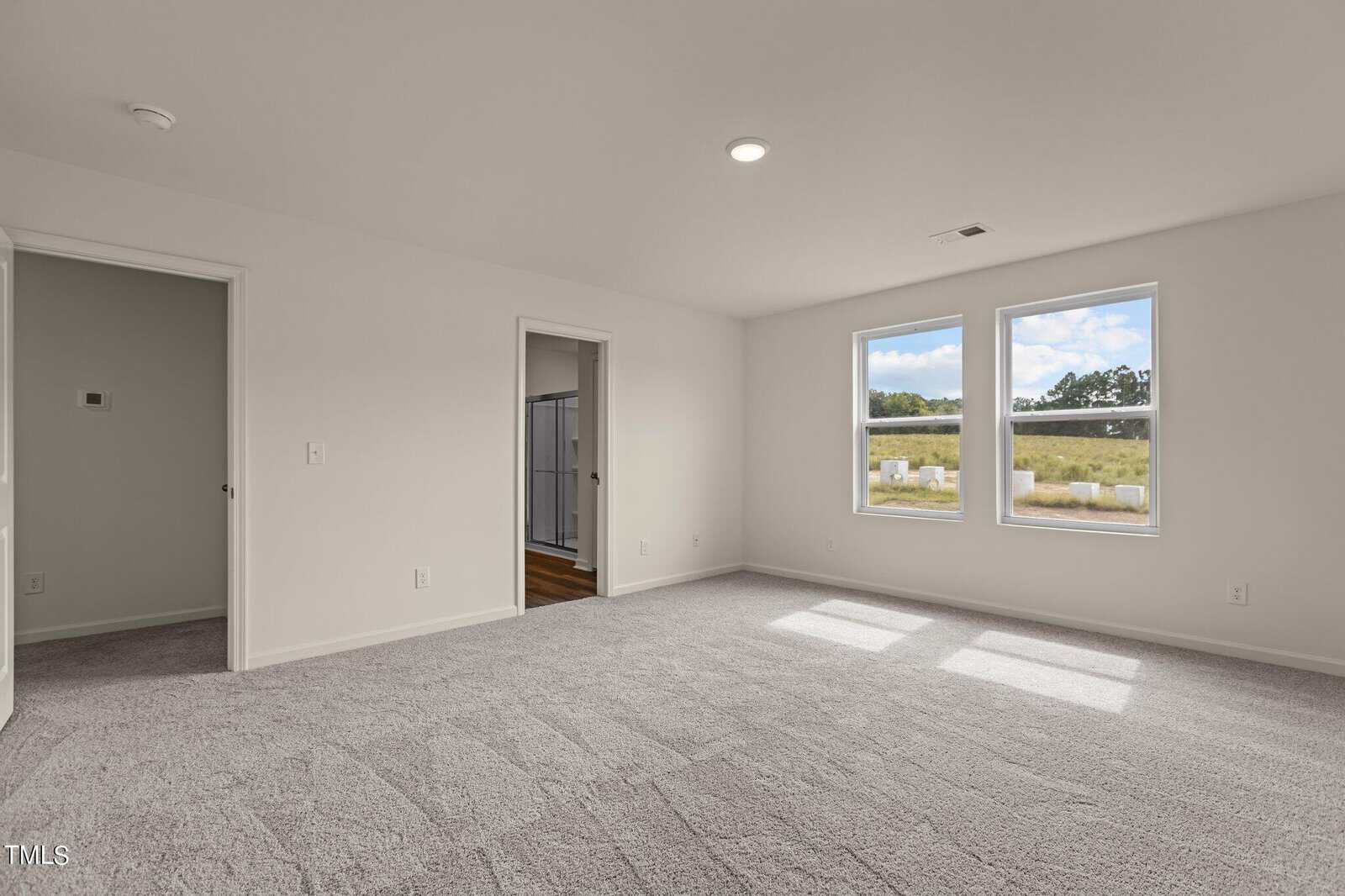 Spacious empty bedroom with light gray carpet, white walls, two windows showing a grassy outdoor view, and doorways leading to an adjacent room and bathroom.