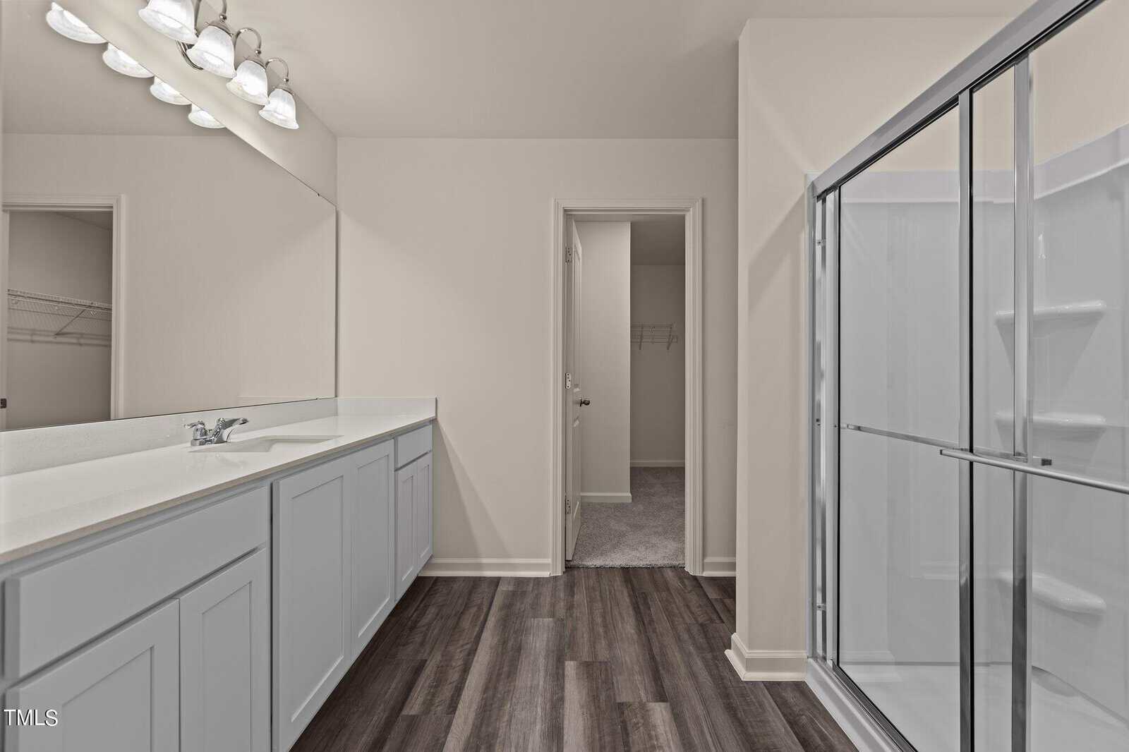Modern bathroom with double sink vanity, large mirror, glass door shower, white cabinets, and wood-look flooring. A door leads to a carpeted closet area in the background.