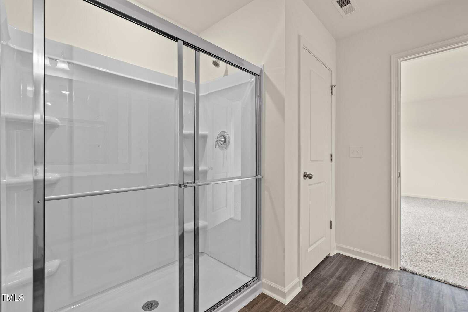 A modern bathroom with a glass-enclosed shower, white walls, dark wood flooring, and a door leading to a carpeted room.