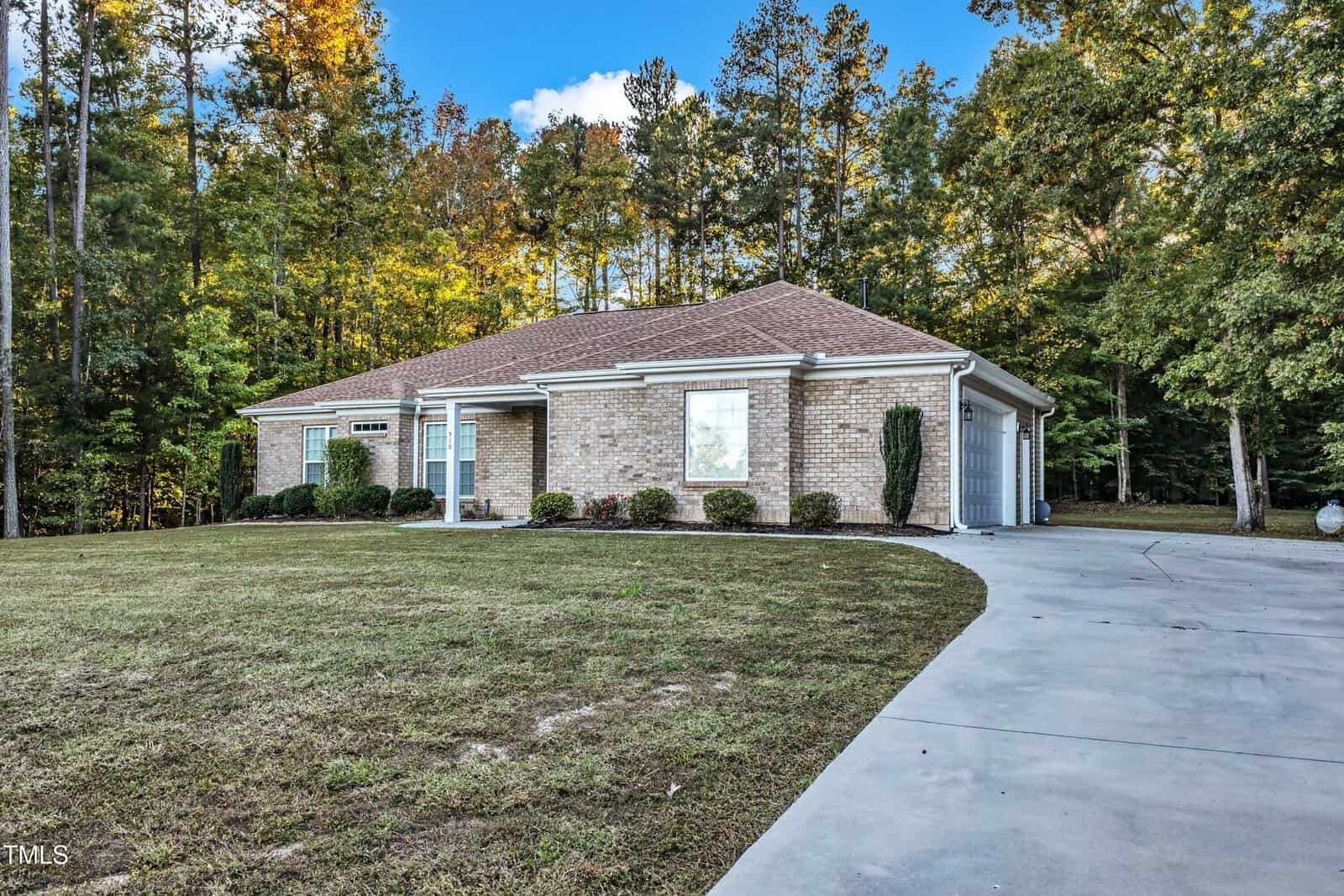The brick single-story house at 510 Willow Creek Run in Henderson boasts a sloped shingle roof and is nestled among trees. It features a wide driveway, a well-maintained lawn with minimal shrubbery, and a clear blue sky with scattered clouds in the background.