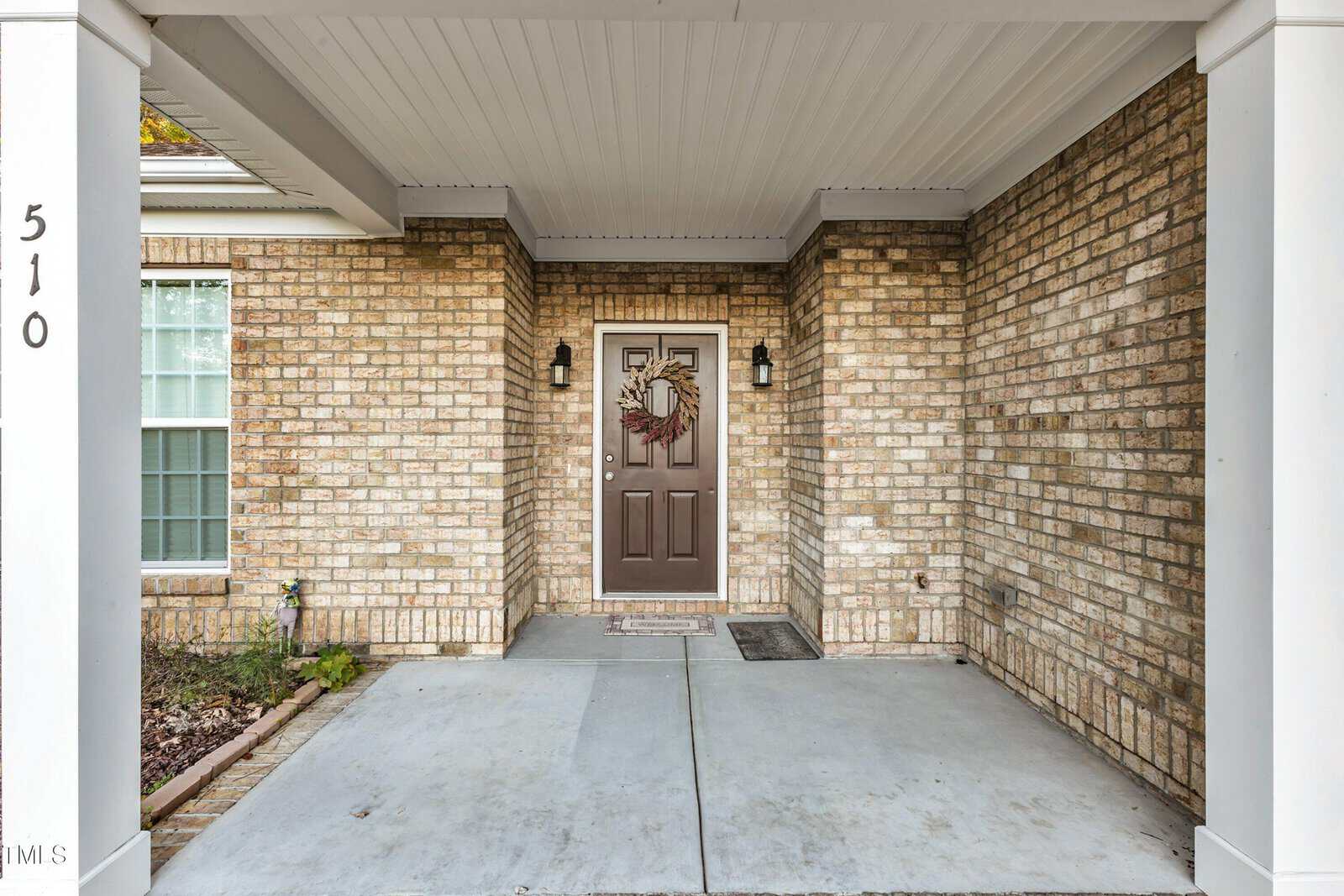 Front entryway of a house with a brown door adorned with a wreath. The exterior features beige brick walls and two black wall lanterns flanking the door. The number 510 is displayed vertically on the left. A small doormat is placed in front of the door.