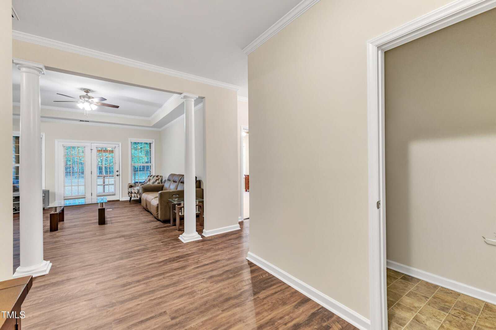 A modern living room with wooden flooring and cream walls. There's a ceiling fan, a sofa, and a glass coffee table. Large windows and a glass door lead to an outside deck. A hallway on the right features a tiled floor.