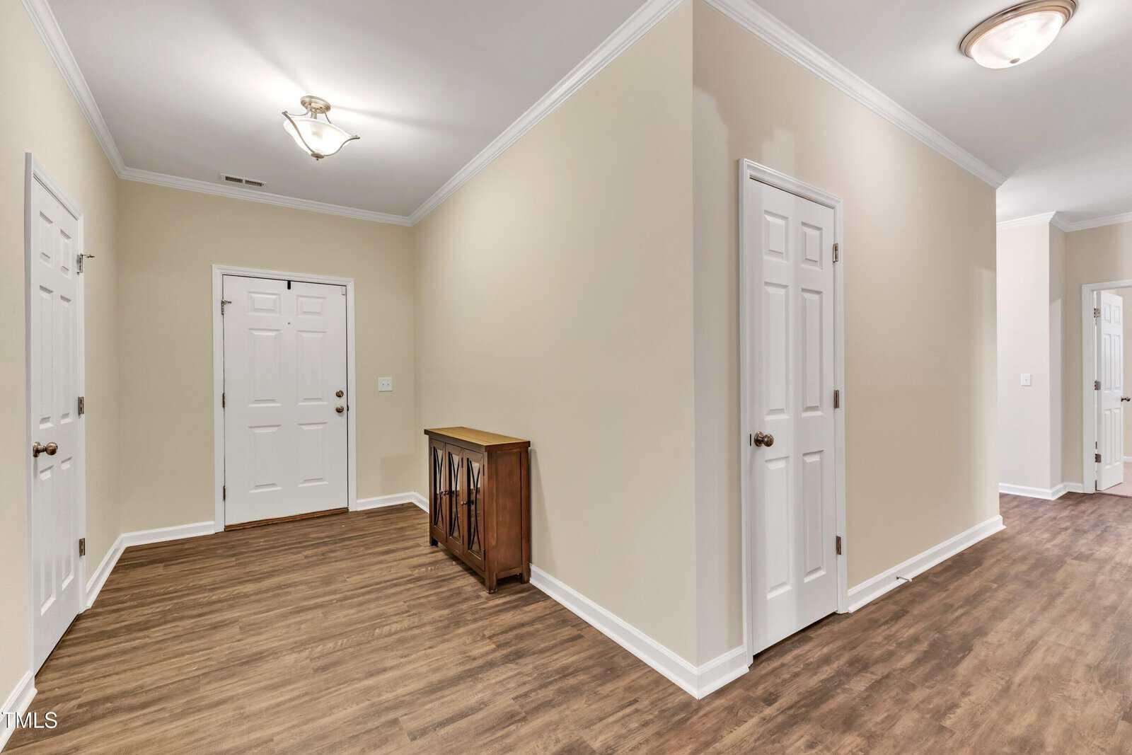 A hallway with beige walls and wood flooring. It features three white doors with dark handles. A small wooden cabinet is against a wall. The area is lit by two ceiling lights.