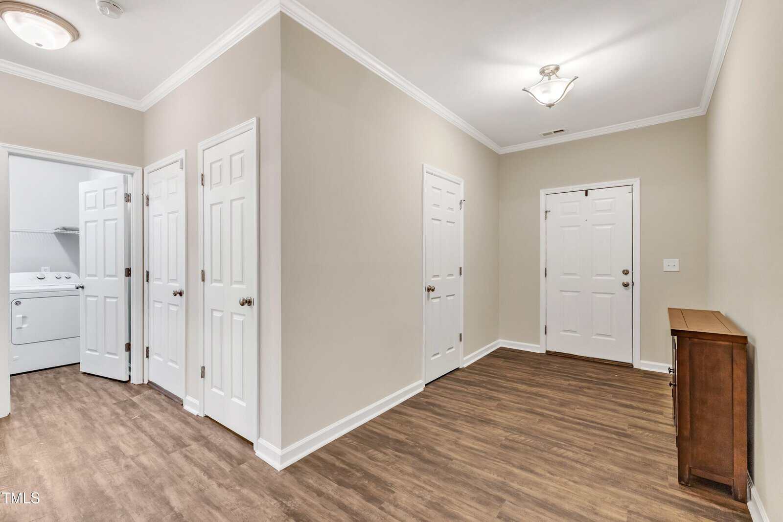 Bright hallway with wood flooring, light beige walls, and white doors. Includes a visible laundry area with a washer and dryer to the left. A small wooden cabinet stands against the right wall. Ceiling light fixture provides illumination.