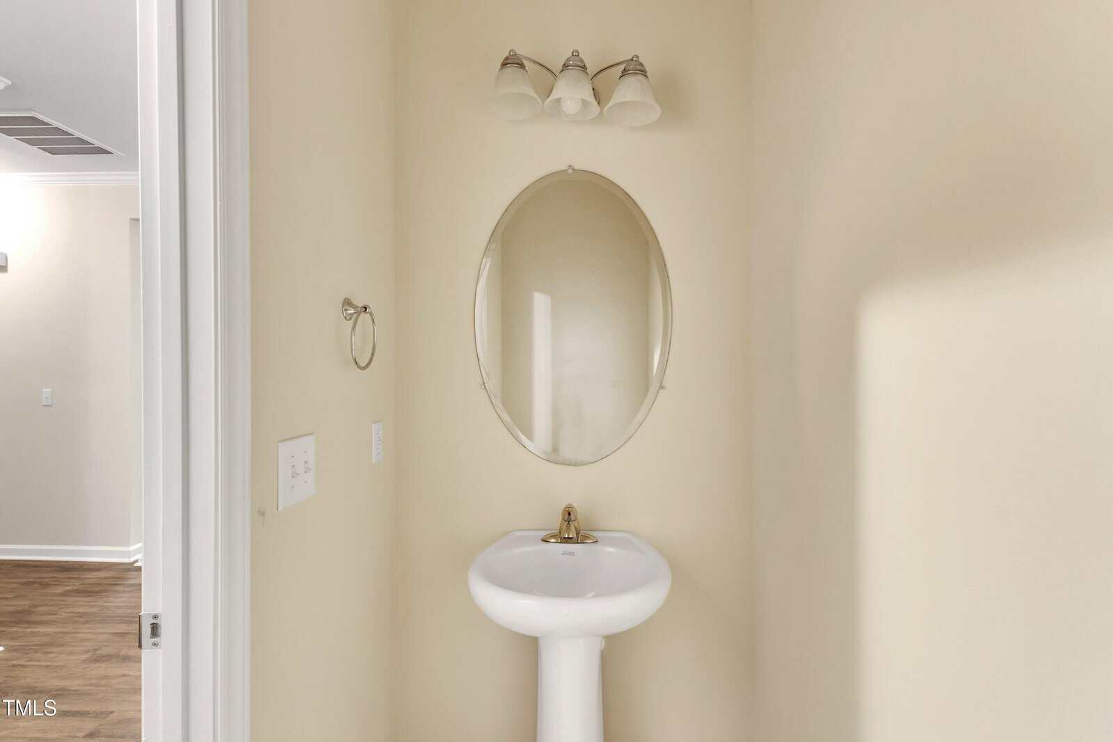 A small bathroom with beige walls featuring an oval mirror above a white pedestal sink. Three-light fixture above the mirror. A towel ring is on the wall to the left, and the floor is wood-toned.