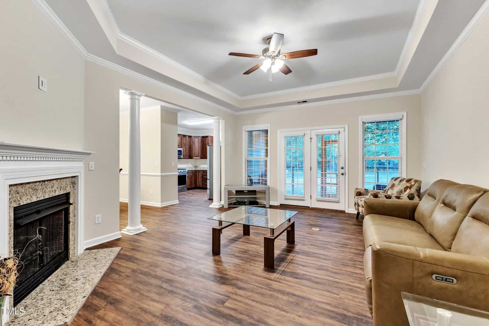 A cozy living room with beige walls and a wooden floor features a ceiling fan and recessed lighting. It includes a tan leather sofa, a glass coffee table, and a fireplace. Large windows and doors open to an outdoor view. A kitchen is visible in the background.