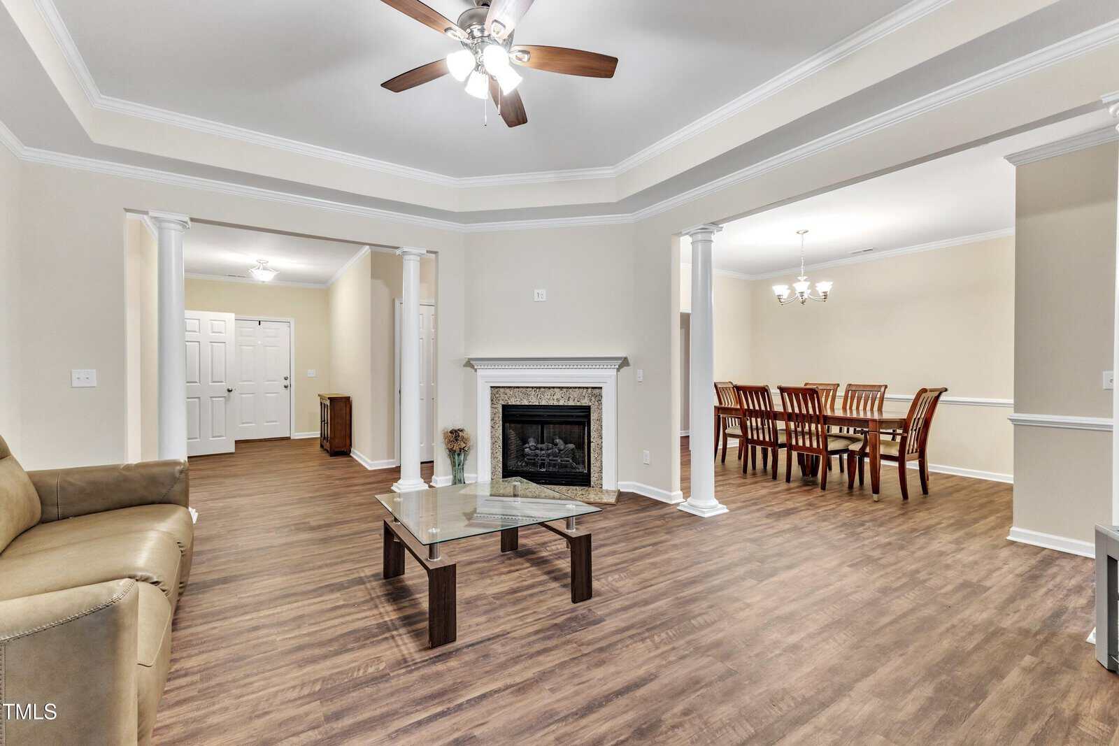 Spacious living and dining area with a light brown sofa, a glass coffee table, a fireplace, and a wooden dining table with chairs. Wood flooring and ceiling fan add warmth to the bright, open-concept space.