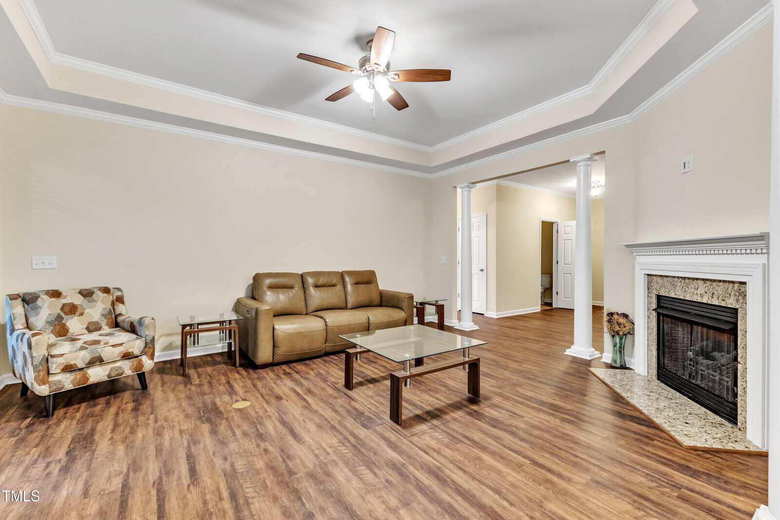 A spacious living room with wooden flooring, featuring a brown leather sofa and patterned armchair. A glass coffee table is centered in front of a fireplace with a mantel. A ceiling fan is overhead, and columns frame an open entryway to another room.