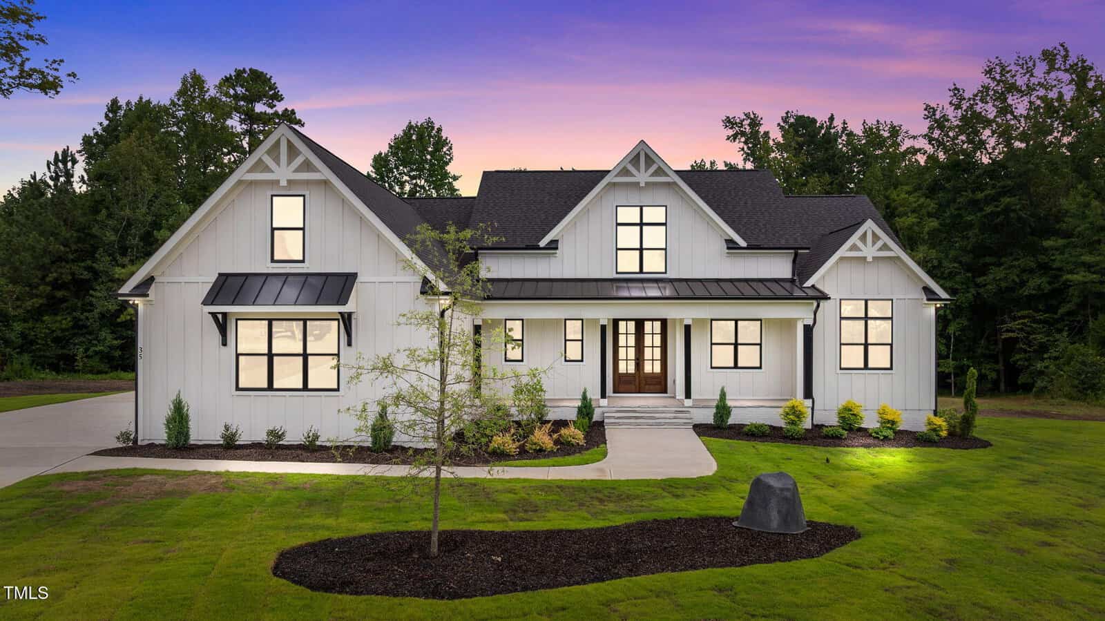 A modern two-story white house with black trim, a dark roof, and large windows, sits on a well-maintained lawn with minimal landscaping, surrounded by trees at sunset.