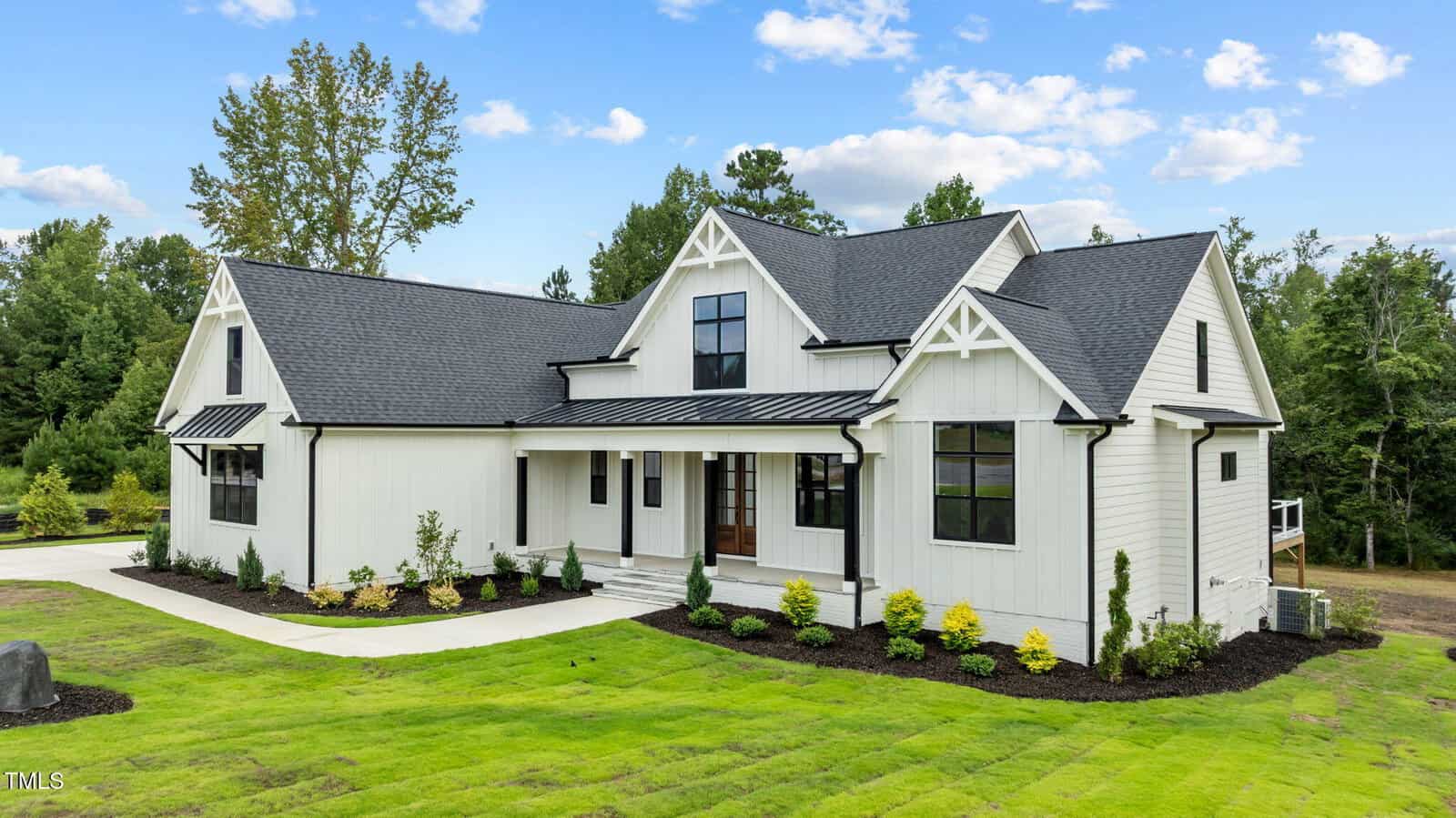 A modern farmhouse-style home with white vertical siding, black trim, peaked roofs, large windows, landscaped front yard, and a concrete driveway, set against a backdrop of green trees and blue sky.