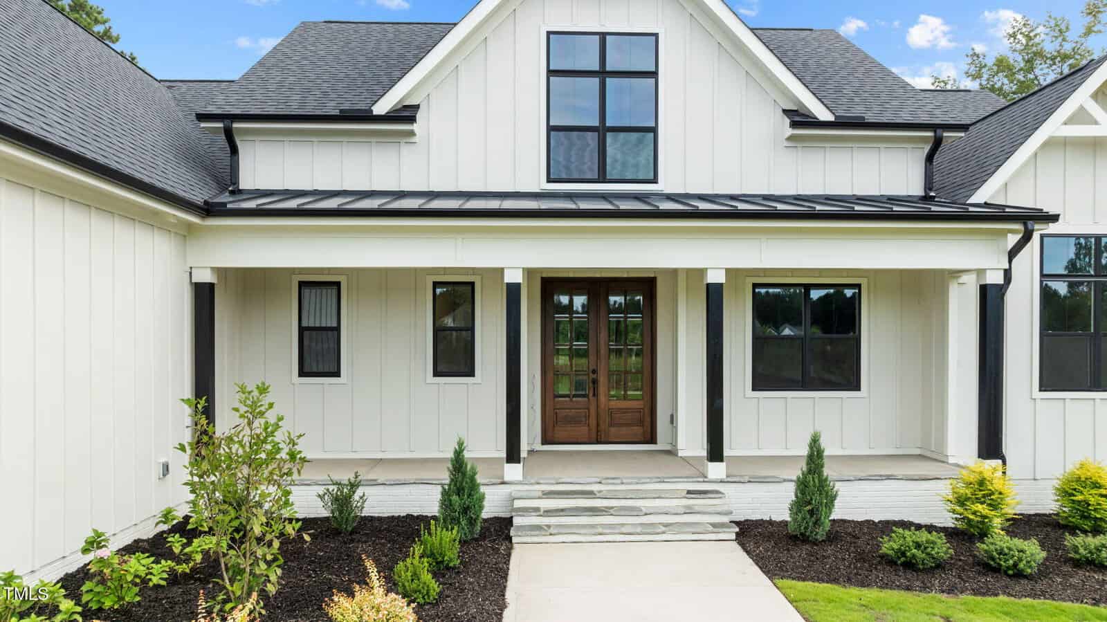 A modern farmhouse with white vertical siding, black trim, a dark metal roof over the porch, double wooden front doors with glass panels, and landscaped shrubs along a concrete walkway.
