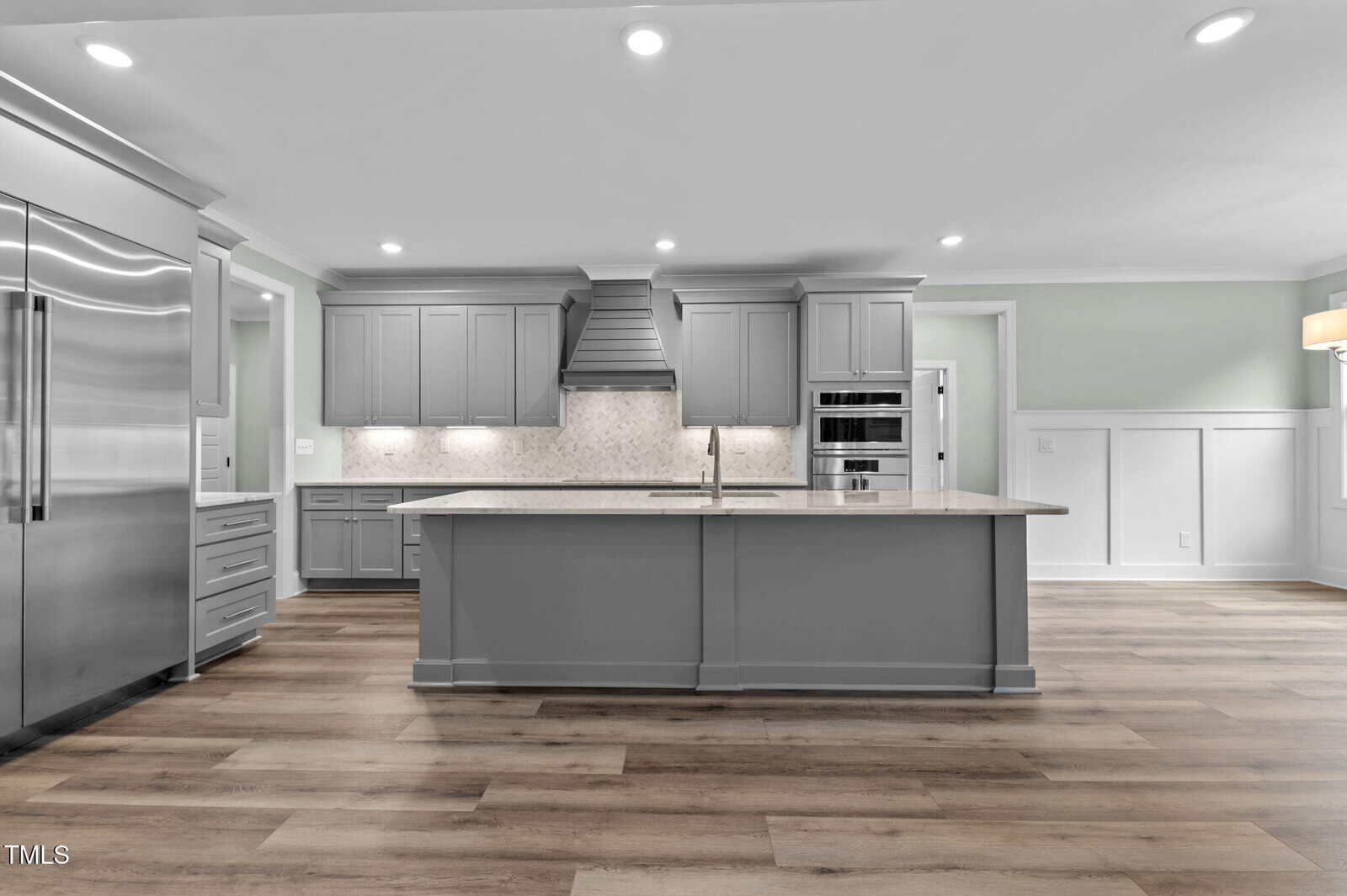 Modern kitchen with gray cabinets, stainless steel appliances, a large island with sink, and light wood flooring. Recessed lighting and a tile backsplash add to the clean, contemporary design.