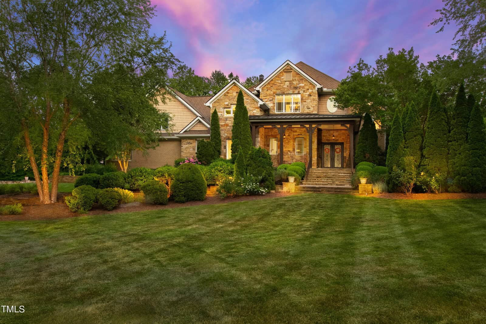 A large stone and brick house with a gabled roof is surrounded by trees and manicured shrubs. It features a front porch with pillars and steps leading to the entrance. The sky is pink and purple at sunset, and the lawn is neatly groomed.