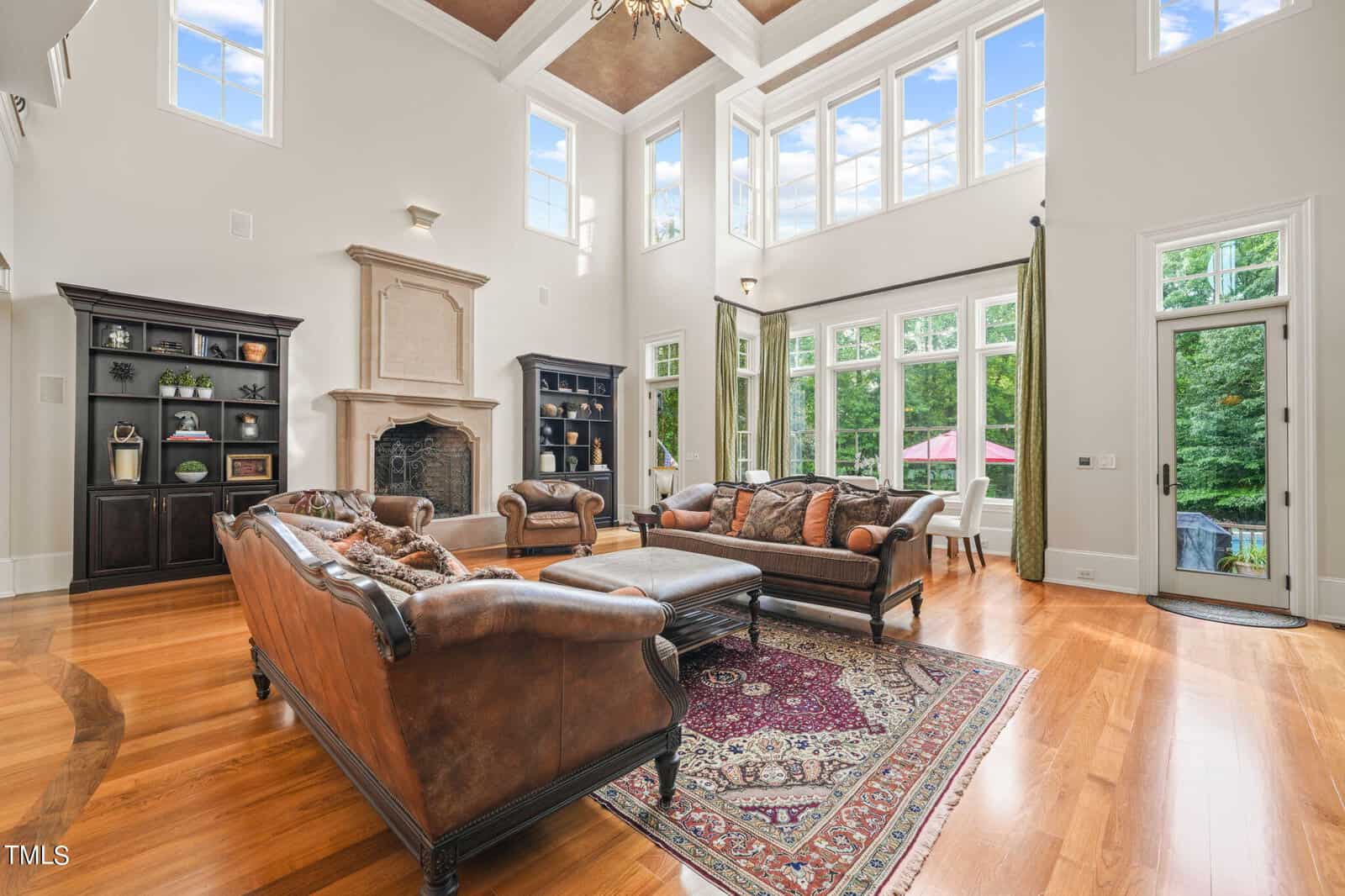 Spacious living room at 1076 Cabin Creek Road in Oxford, featuring high ceilings and large windows. A decorative fireplace complements the leather sofas and patterned rug. Shelves and a small dining area are visible, with hardwood floors and an outside view of greenery completing the setting.