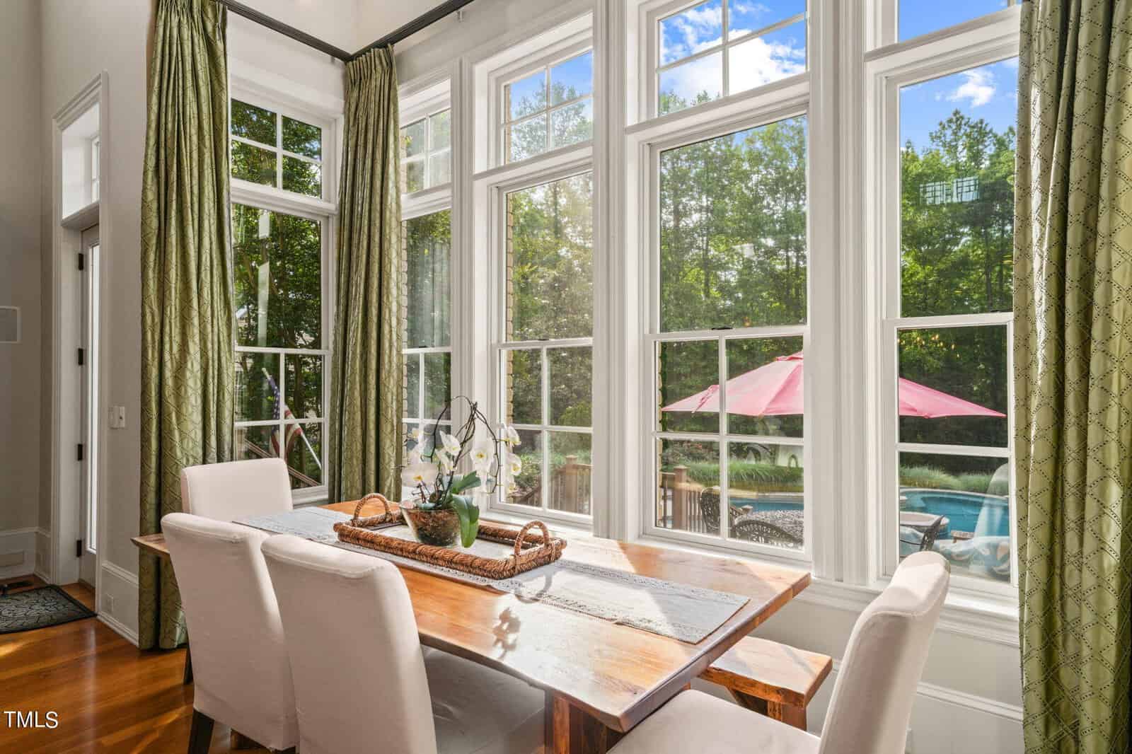 Elegant dining area with a wooden table and white cushioned chairs next to large windows. Outside, there's a view of a deck with a pink umbrella and a pool surrounded by trees. Green curtains adorn the tall windows, and sunlight brightens the room.
