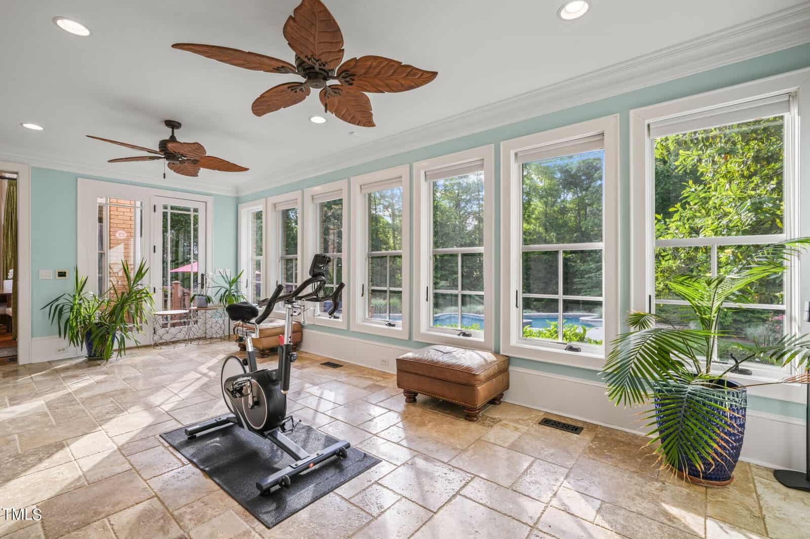 A bright room with large windows showing a greenery view. There is an exercise bike on a mat facing the windows. The room features tile flooring, ceiling fans with leaf-shaped blades, a brown ottoman, and potted plants.