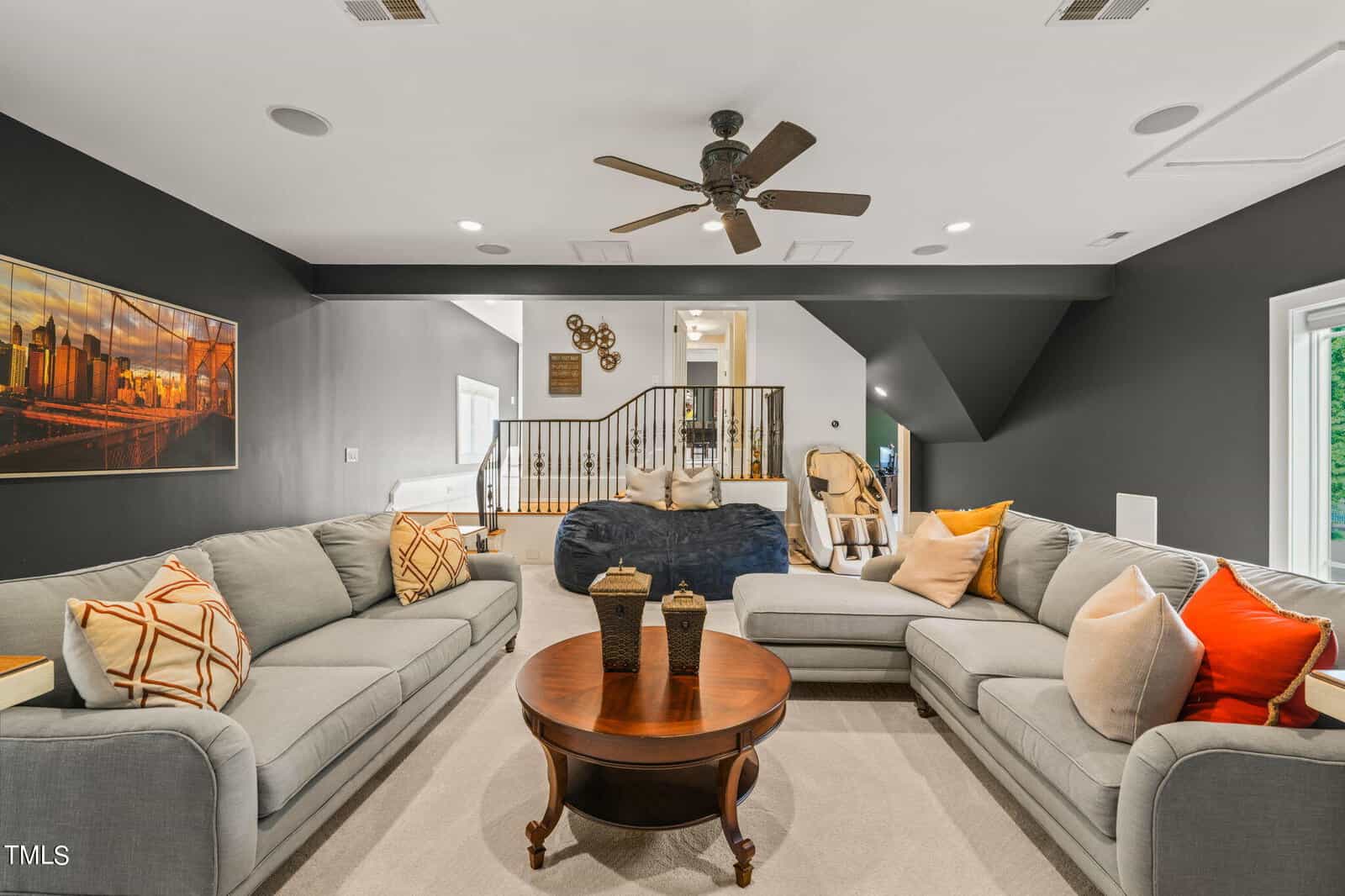 A modern living room with gray walls and a sectional sofa adorned with colorful throw pillows. A round wooden coffee table sits in the center. A ceiling fan hangs above, and a staircase with a wooden railing leads to an upper level.