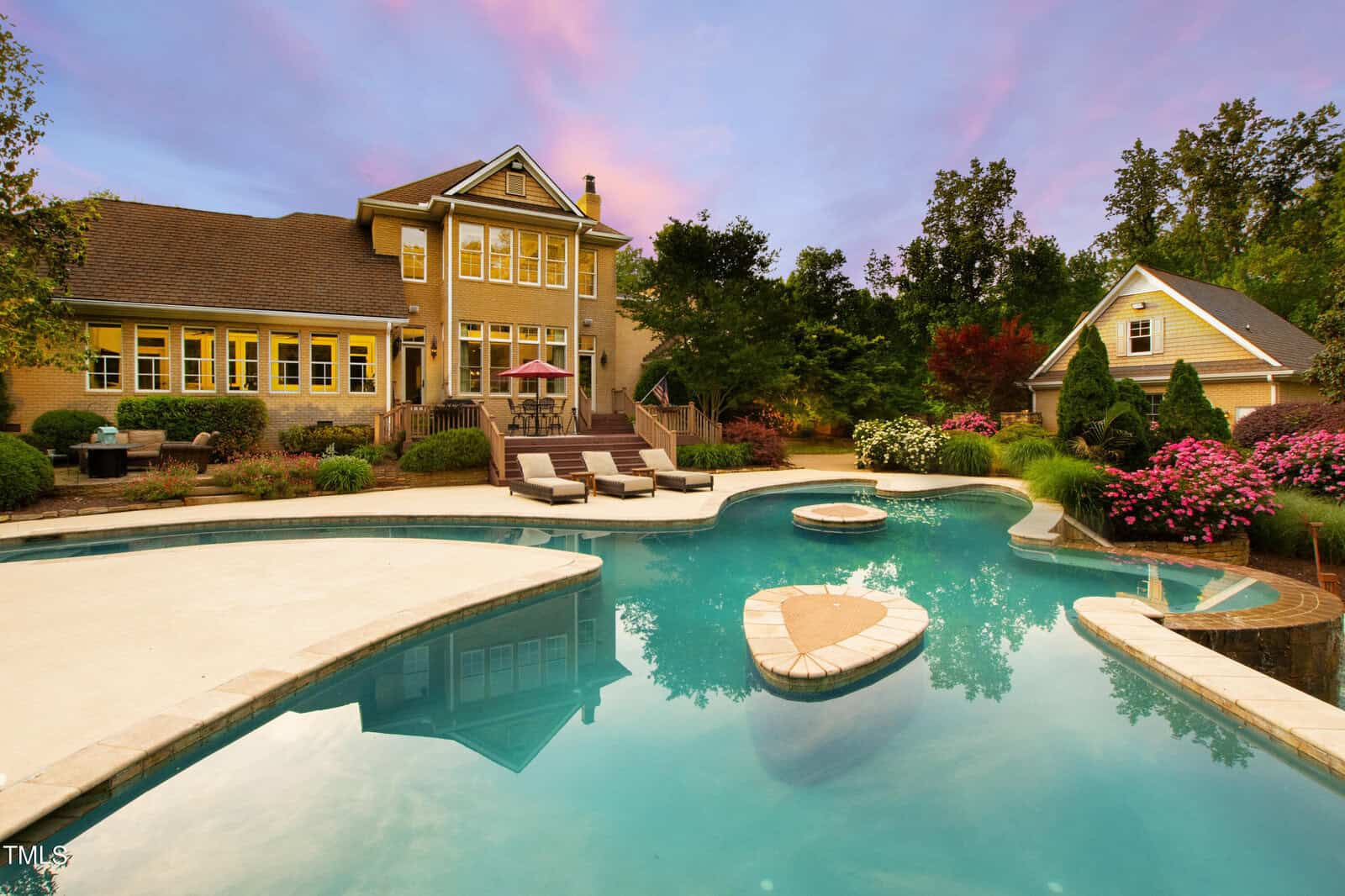 A large backyard with a swimming pool featuring a small island, surrounded by neatly manicured landscaping and trees. A patio with lounge chairs and an umbrella is adjacent to a two-story house with large windows and light-colored siding.
