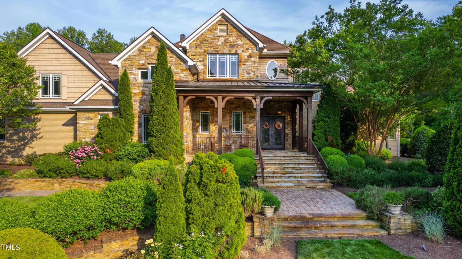 Large stone house with a gabled roof and wooden accents. The entrance features a set of stairs leading to a covered porch, surrounded by manicured shrubs and trees. The facade combines stone and wood siding with decorative elements.