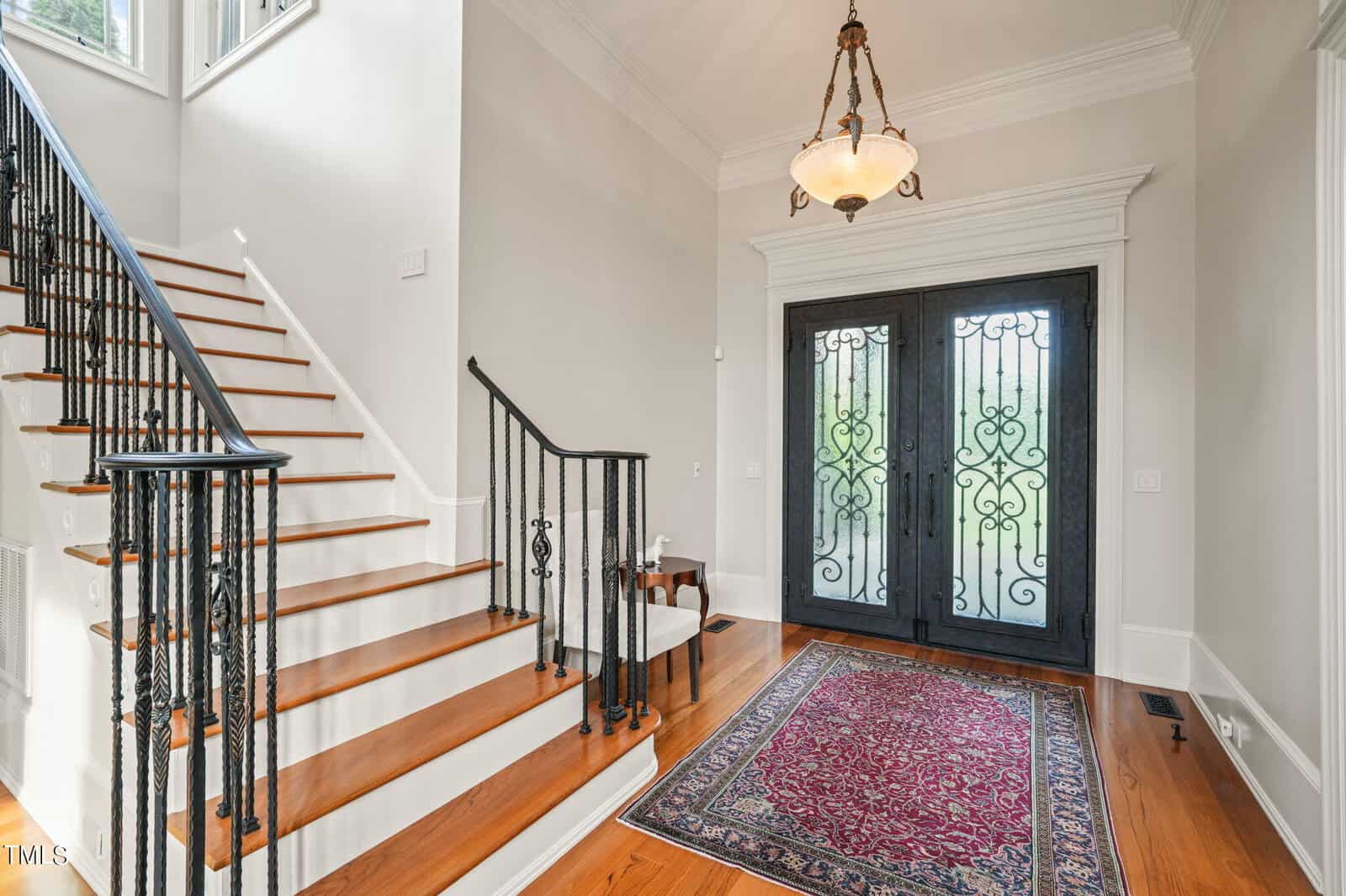 A foyer with a grand iron and glass double door, a staircase with wooden steps and black metal railing on the left. A red patterned rug lies on the wooden floor. An elegant chandelier hangs from the ceiling, and light streams through large windows.