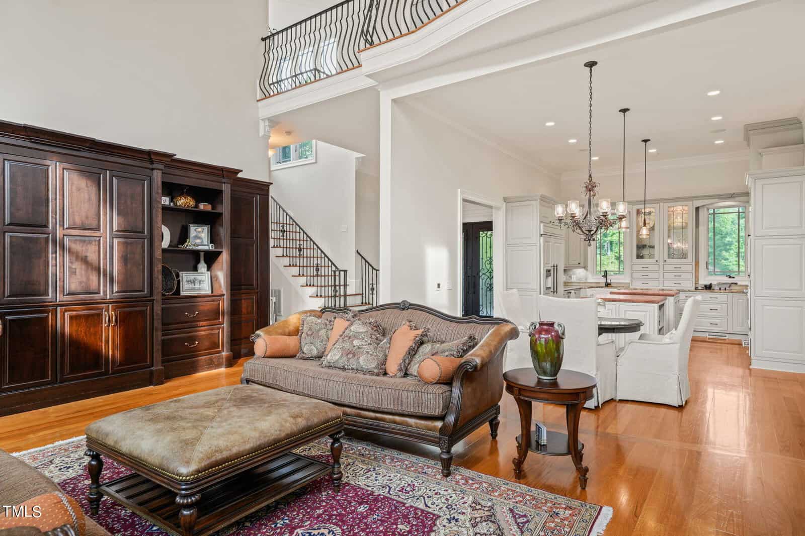 A spacious living room with a brown leather sofa and matching ottoman on a patterned rug. An ornate chandelier hangs above a dining area in the background. A dark wooden armoire is against the wall, and a staircase leads to an upper floor.
