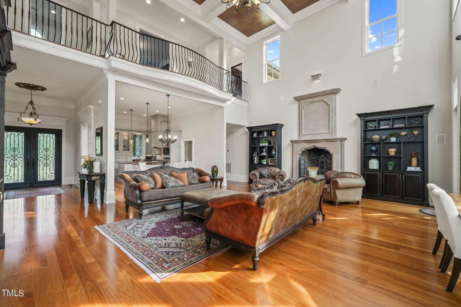 Spacious living room with high ceilings and hardwood floors featuring leather sofas, a wooden coffee table, bookcases, and a fireplace. A decorative rug is on the floor. A balcony overlooks the open layout with a view of the kitchen and front entrance.
