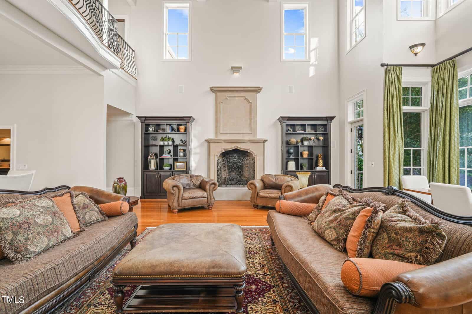 Located at 1076 Cabin Creek Road in Oxford, this spacious living room boasts high ceilings and large windows. It features two brown sofas with patterned cushions, a large ottoman, and two armchairs. A stone fireplace is flanked by dark bookshelves, with hardwood floors and a patterned rug completing the scene.
